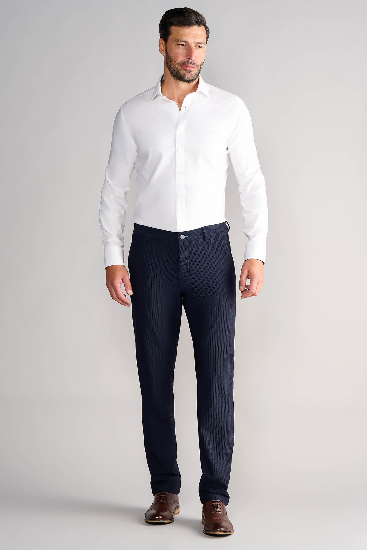 A man with short brown hair and a trimmed beard stands against a light gray background, wearing a white dress shirt and Twillory AIR Pants in navy, made from performance fabric with 4-way stretch. His hands rest by his sides.