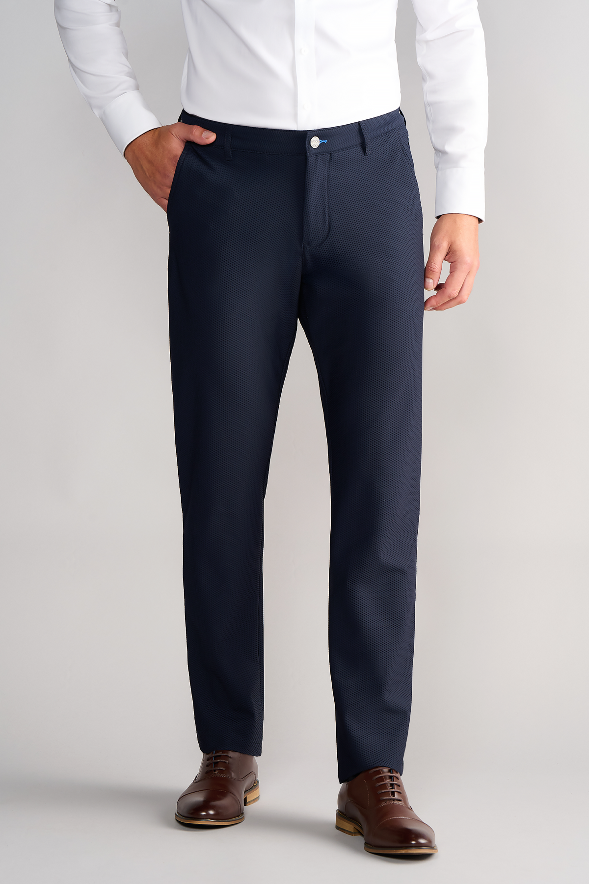 A man, face not visible, stands against a plain background wearing a white shirt tucked into Twillory AIR Pants in Navy—crafted from performance fabric—with one hand in his pocket and paired with brown leather dress shoes.