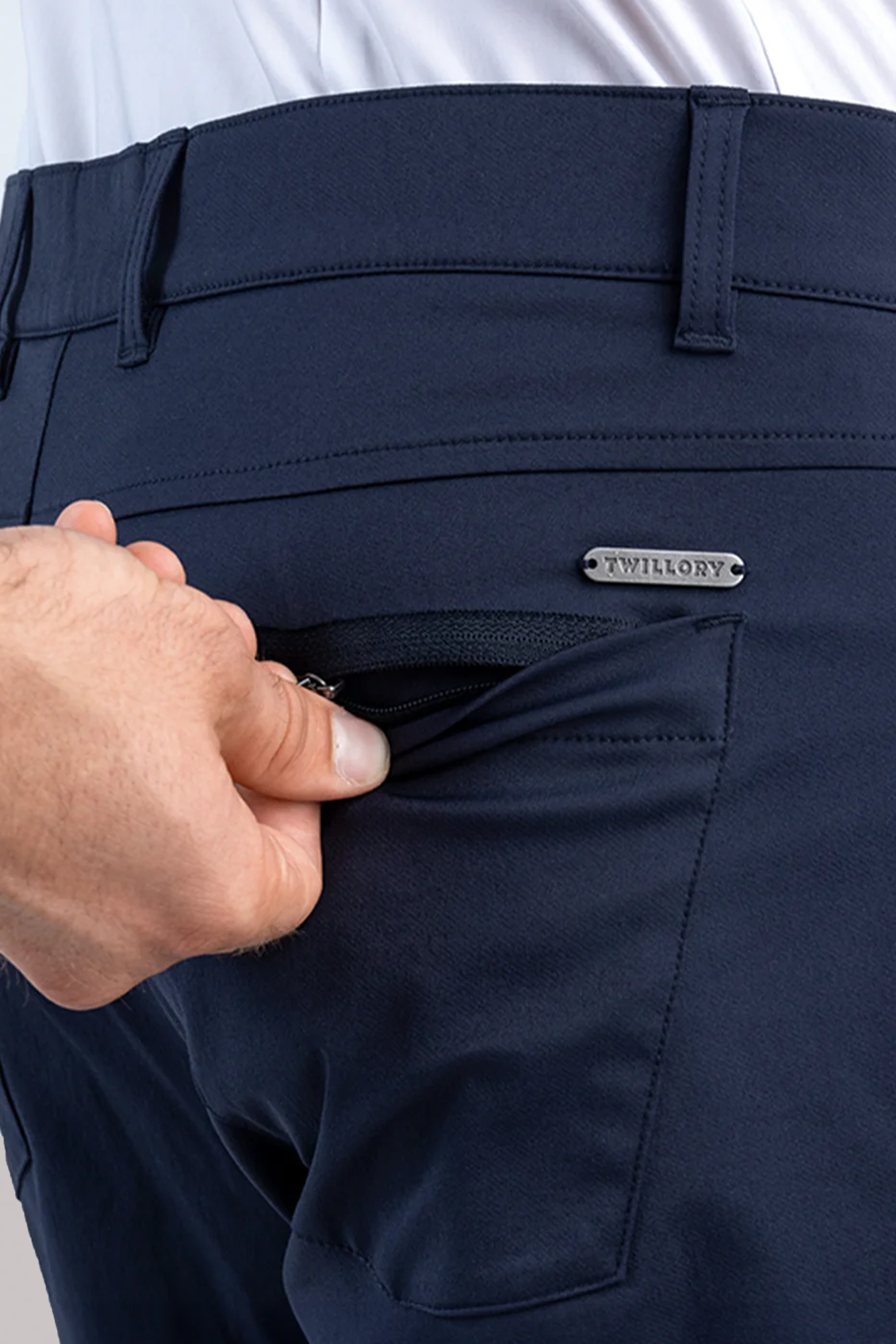 A close-up of a hand opening the zippered back pocket on Twillory Performance Five Pocket // NAVY pants, featuring belt loops and a metallic Twillory label above the pocket. The zipper pull and part of the pocket interior are visible.
