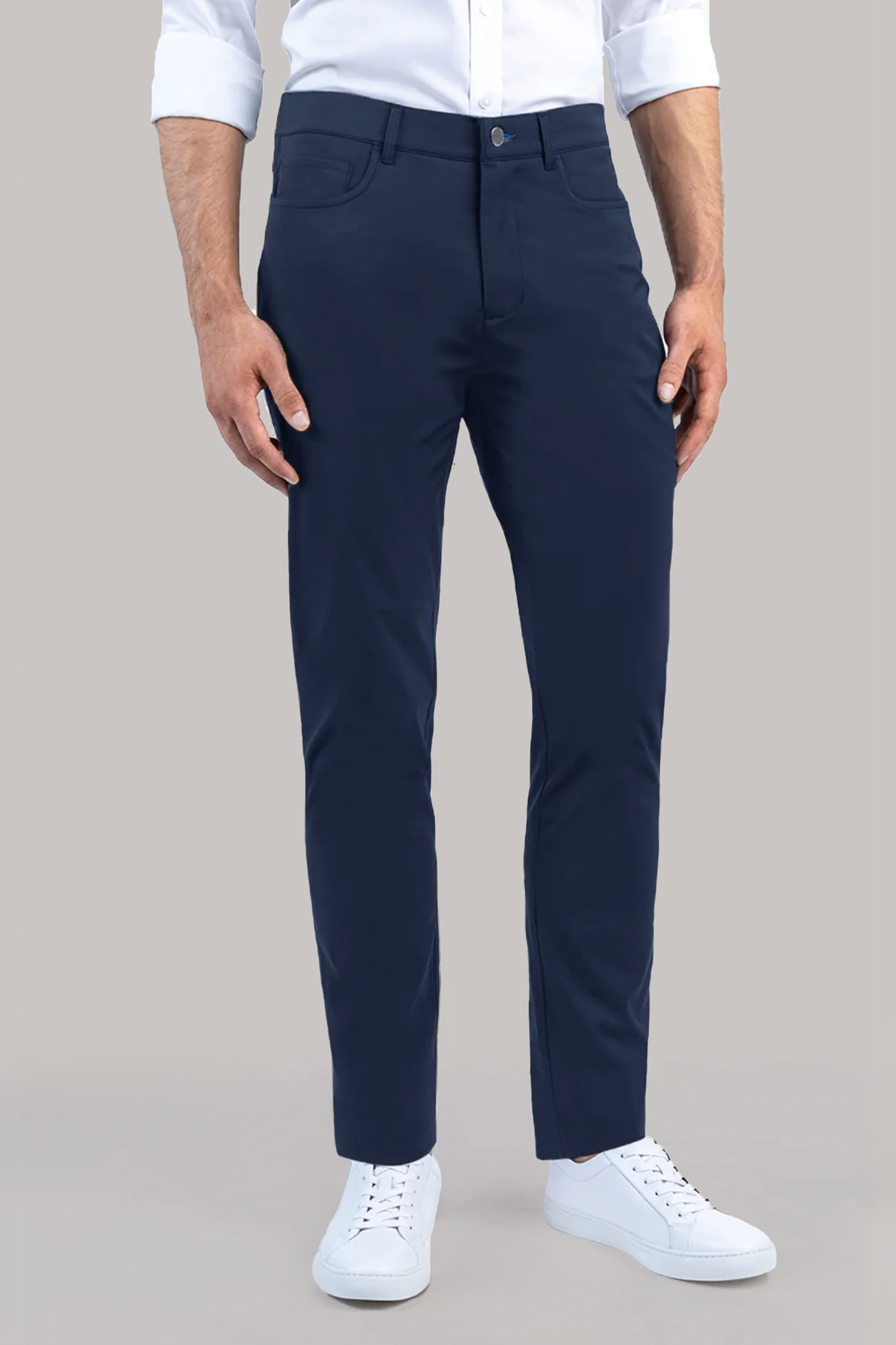 A man stands against a plain light background, wearing Twillory’s Performance Five Pocket // NAVY pants, a white non-iron shirt, and white low-top sneakers. His left hand is by his side and his right touches his thigh; his head is not shown.