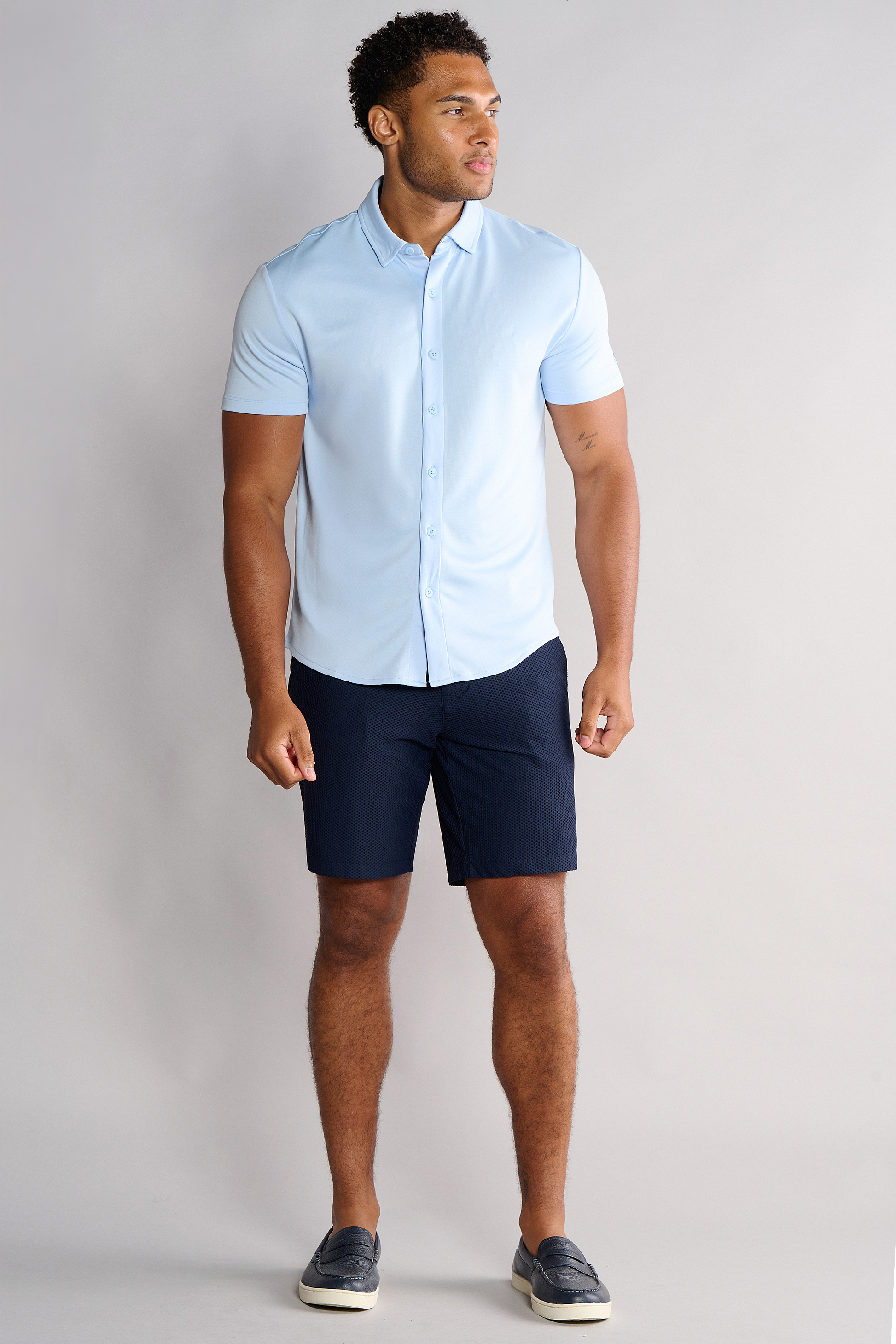 A man stands against a plain light gray background, wearing a light blue short-sleeve button-up shirt, Twillory AIR Shorts, and dark slip-on shoes. He looks slightly to the side with his left hand relaxed and his right hand by his hip.