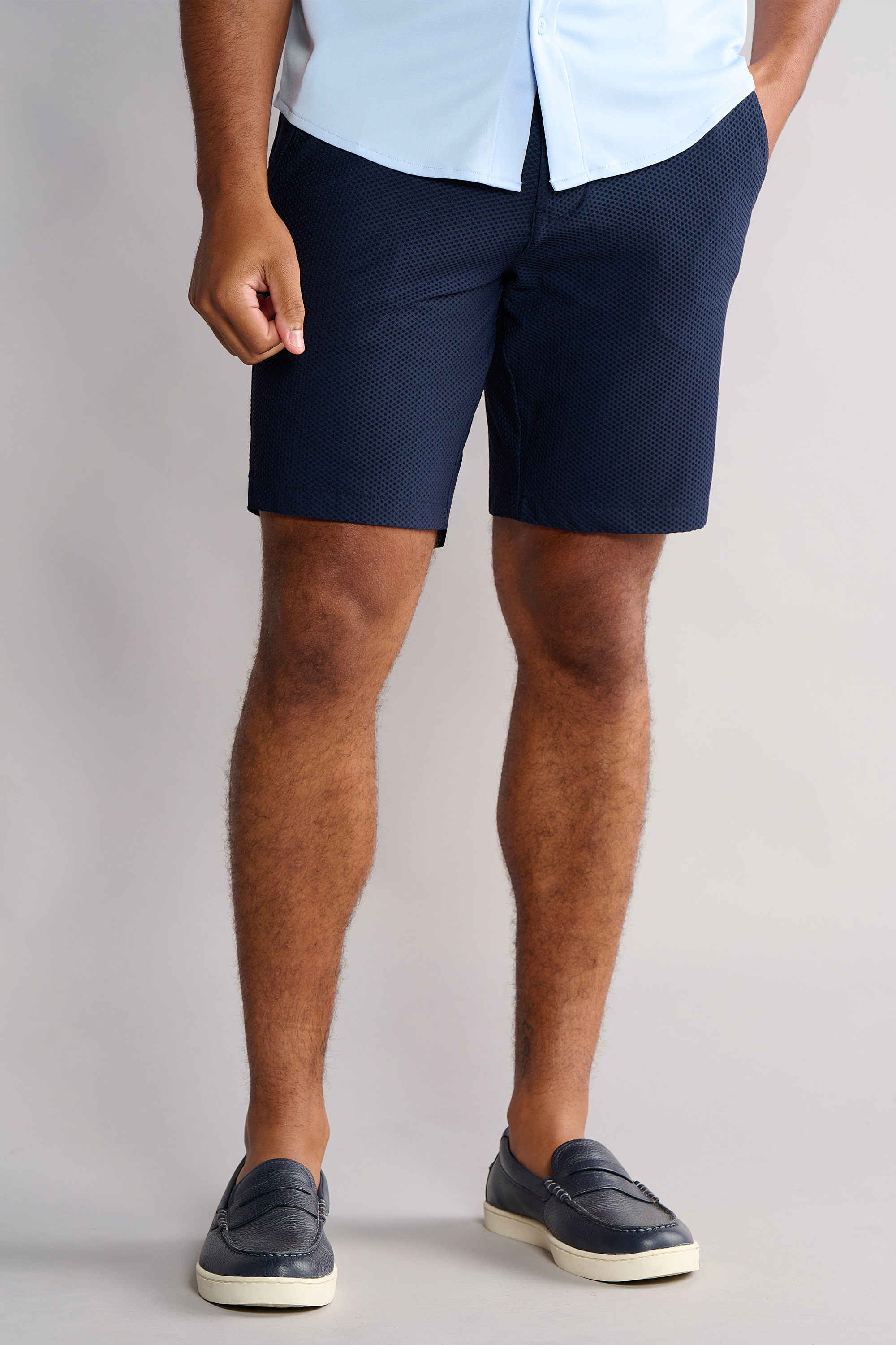 A person stands against a plain background wearing a light blue button-down shirt and navy Twillory AIR Shorts with sweat-wicking technology, paired with navy loafers. Their right hand is in one pocket, and only their body from shoulders to feet is visible.