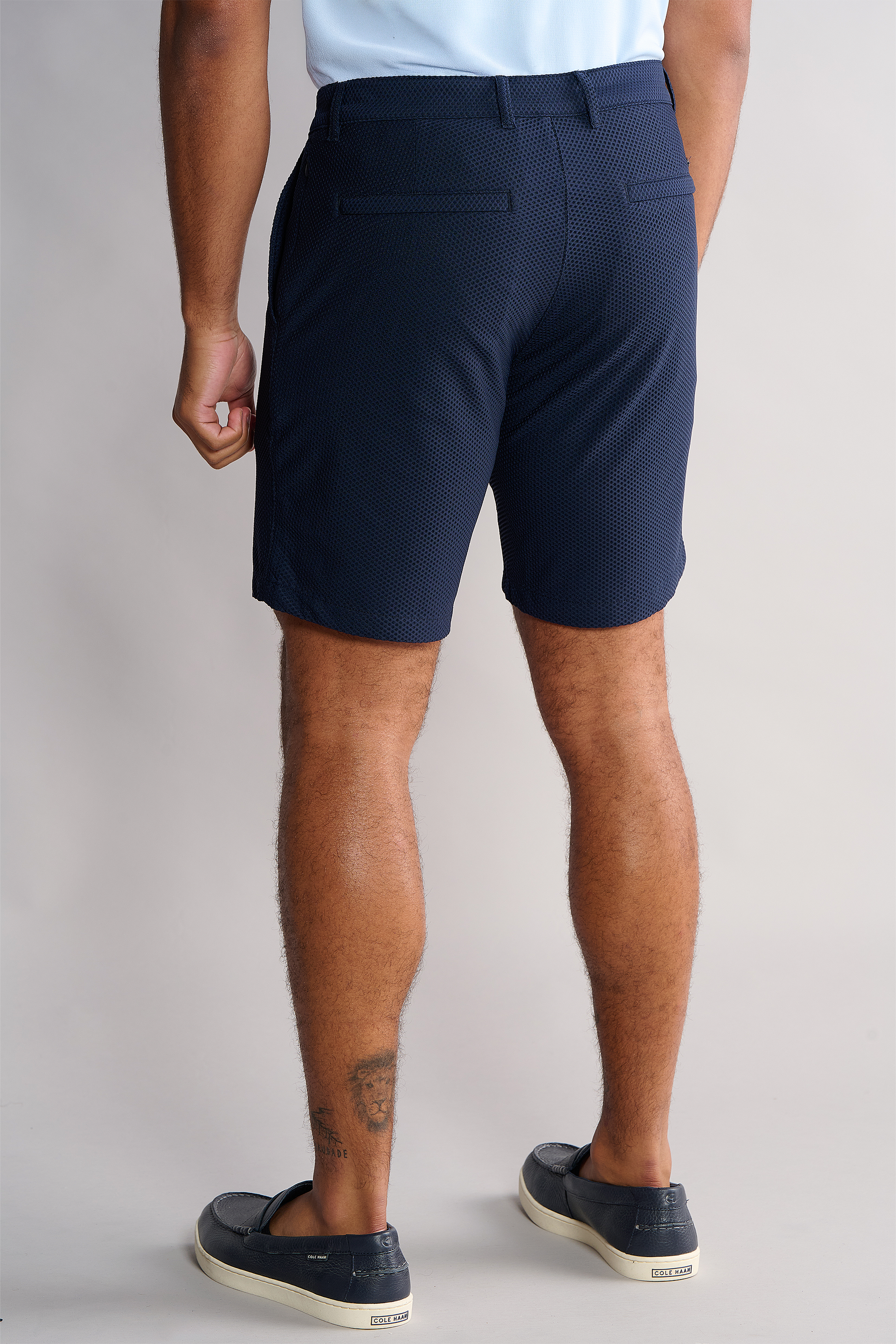 A person is shown from the back, wearing a light blue shirt tucked into Twillory AIR Shorts in dark navy with back welt pockets. They have visible leg hair, a right calf tattoo, and black slip-on shoes with beige soles, against a neutral background.