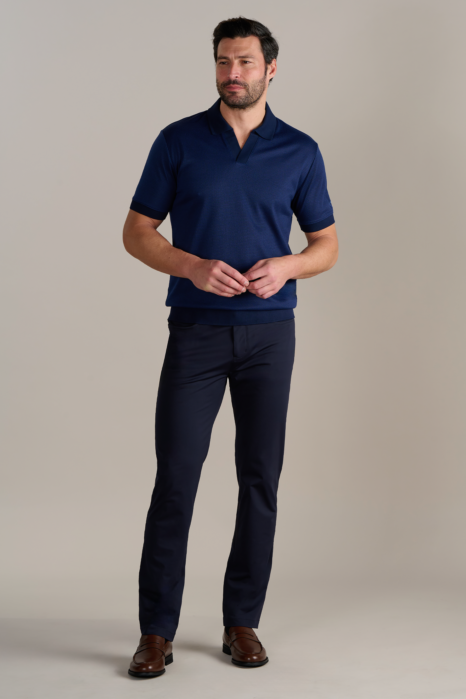 A man with short dark hair and a beard stands against a plain light background, wearing Twillory's Short Sleeve Sweater Polo Solid in Navy, paired with dark slim-fit pants and brown dress shoes. He faces forward, hands loosely clasped at his waist.