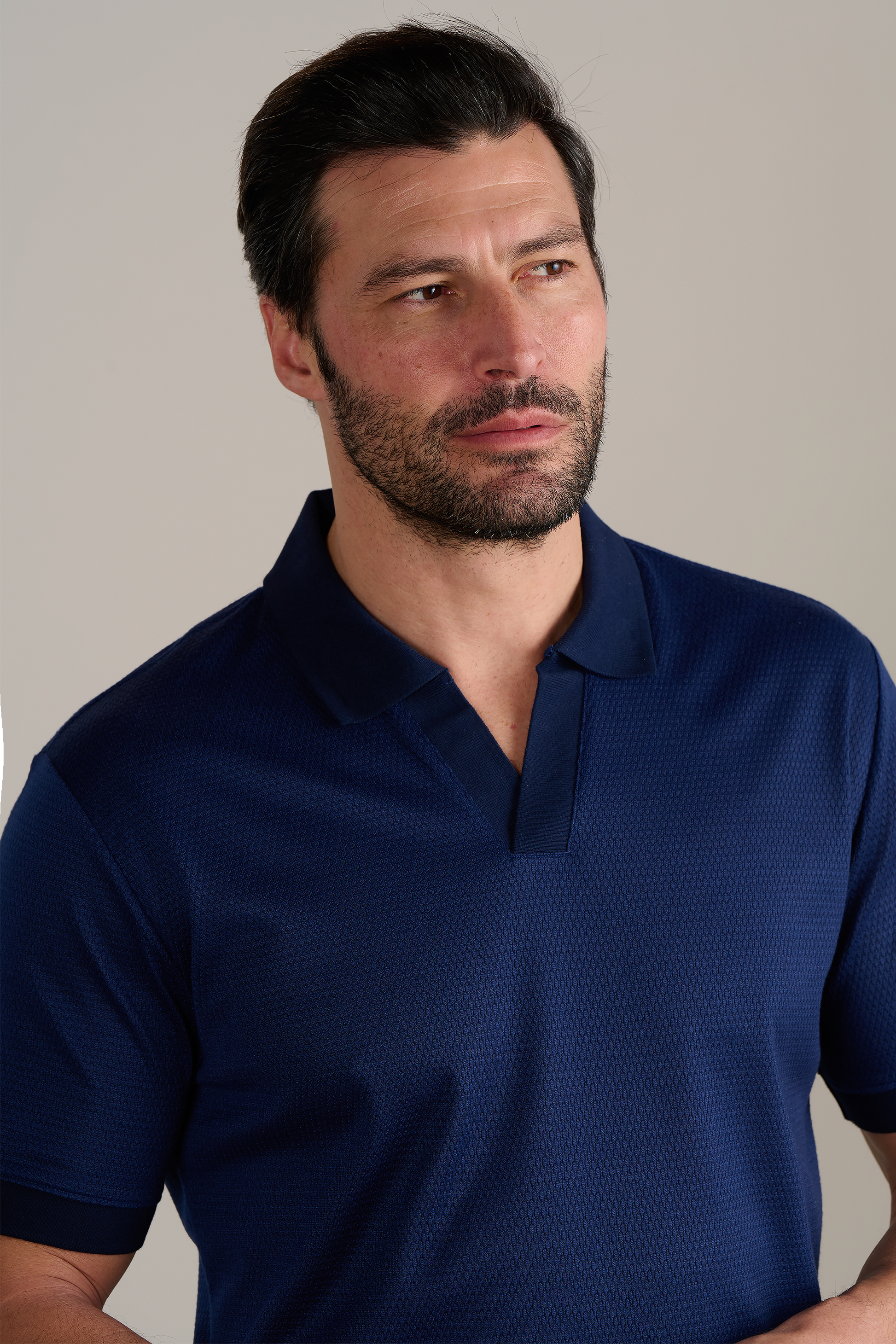 A man with short dark hair and a trimmed beard gazes to his left, wearing the Twillory Short Sleeve Sweater Polo Solid in Navy. The plain, light background highlights his contemplative expression.