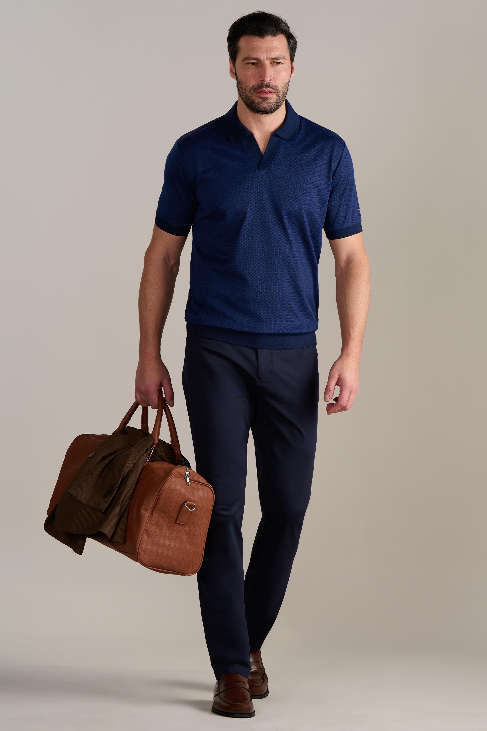 A man with short dark hair and a beard walks forward wearing the Twillory Short Sleeve Sweater Polo Solid in Navy, paired with dark pants and brown loafers. He carries a brown leather duffel bag and has a jacket draped over his arm. The background is plain and light.