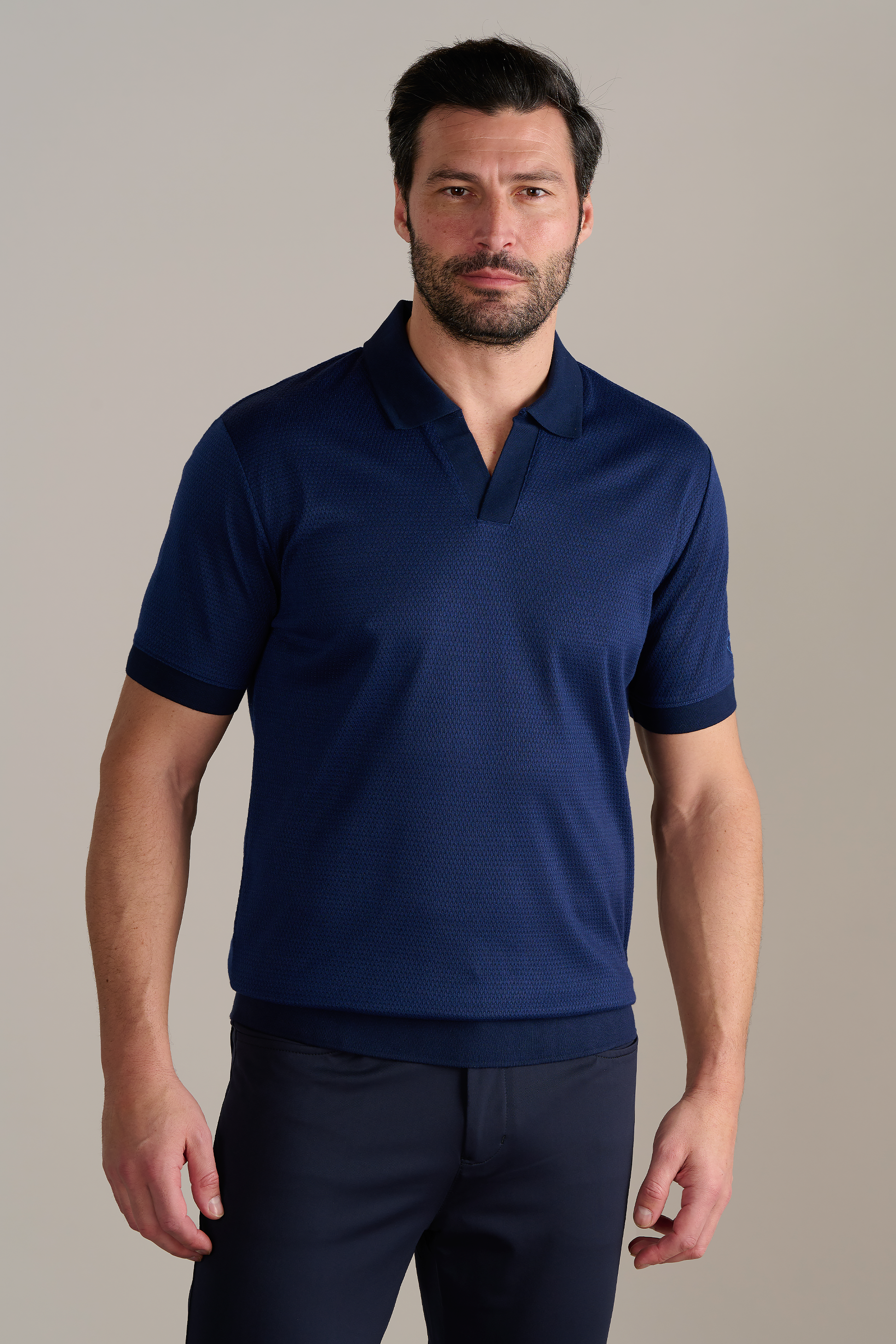 A man with dark hair and a beard wears the Twillory Short Sleeve Sweater Polo Solid in navy with dark pants. He stands against a plain, light background, facing forward with a neutral expression and his arms relaxed at his sides.