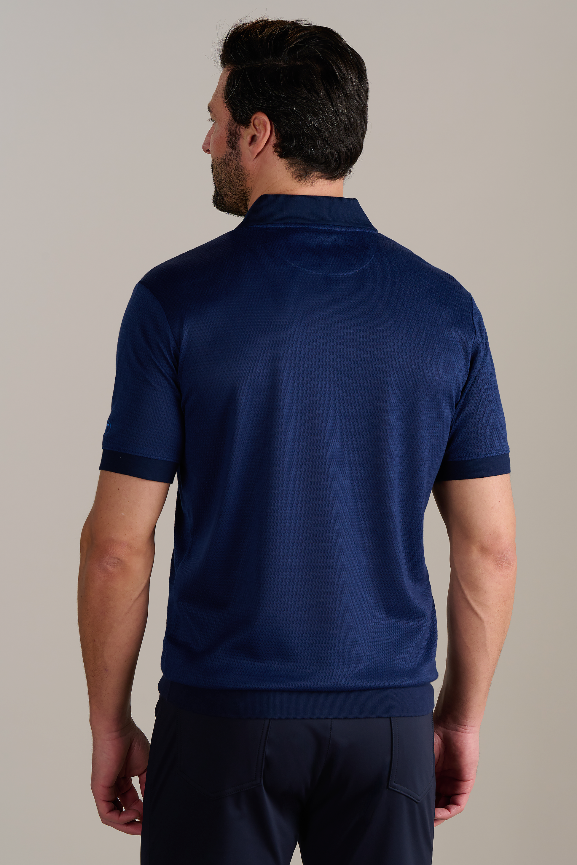 A man with dark hair and a beard stands with his back to the camera, wearing the Twillory Short Sleeve Sweater Polo Solid in Navy. His arms are at his sides against a plain, light background.