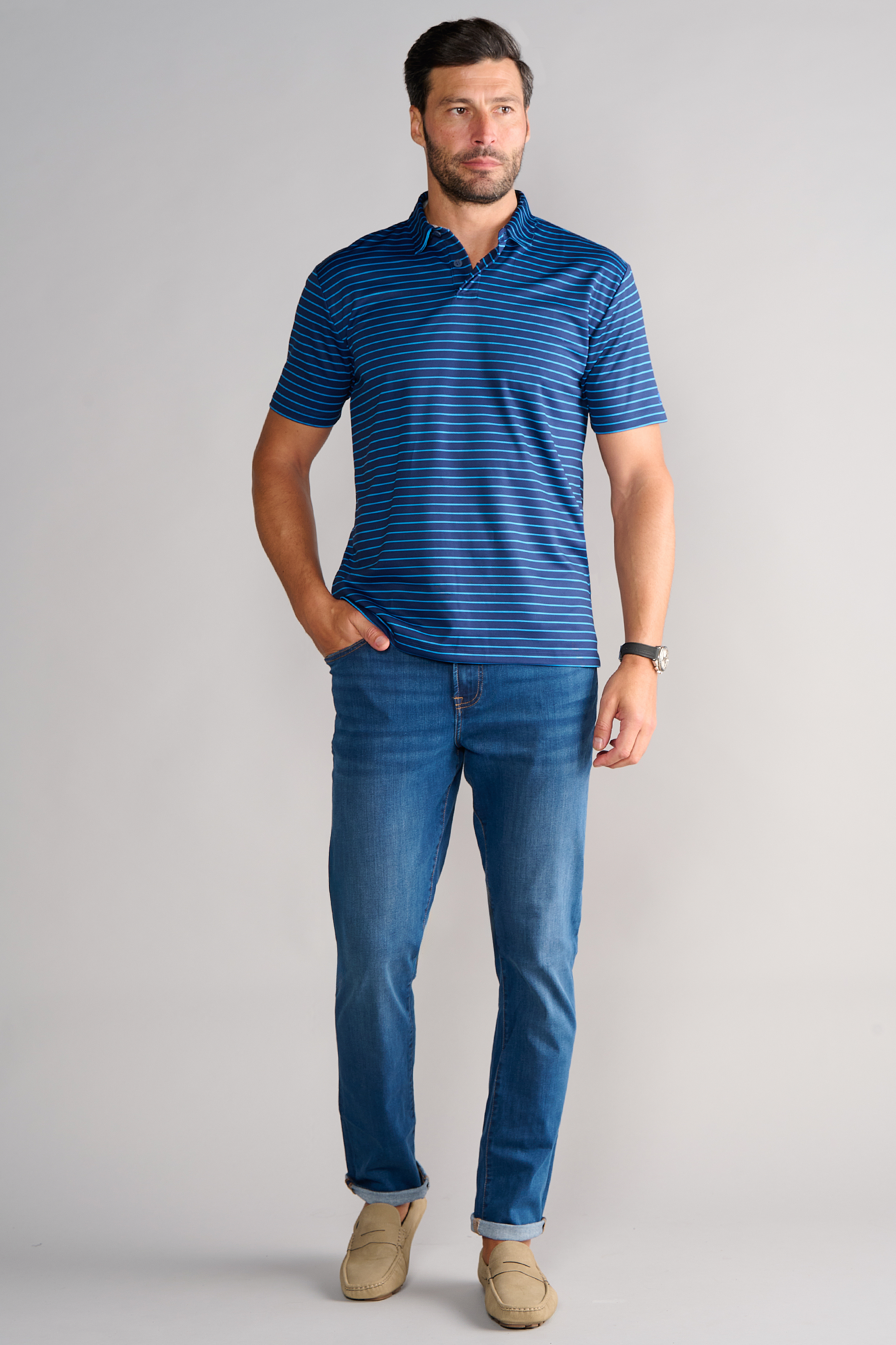 A man with short brown hair and a beard stands against a plain background, wearing the Twillory Short Sleeve Polo Stripe in Navy Blue, paired with blue jeans, beige loafers, and a black watch, with one hand in his pocket.
