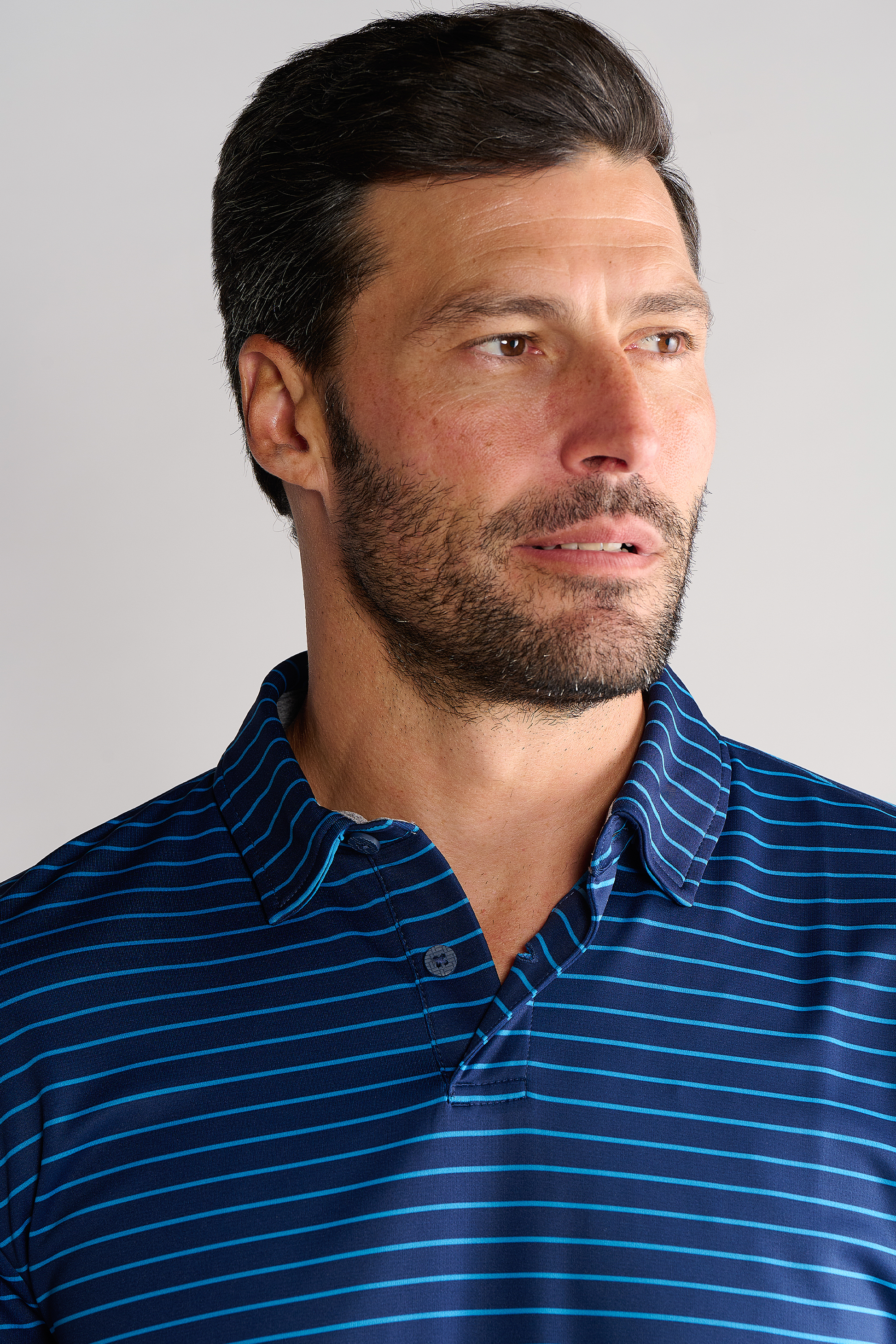 A man with short dark hair and a beard looks to the side, wearing a Twillory Short Sleeve Polo Stripe in navy blue. The background is plain and light-colored, giving him a calm and thoughtful appearance.