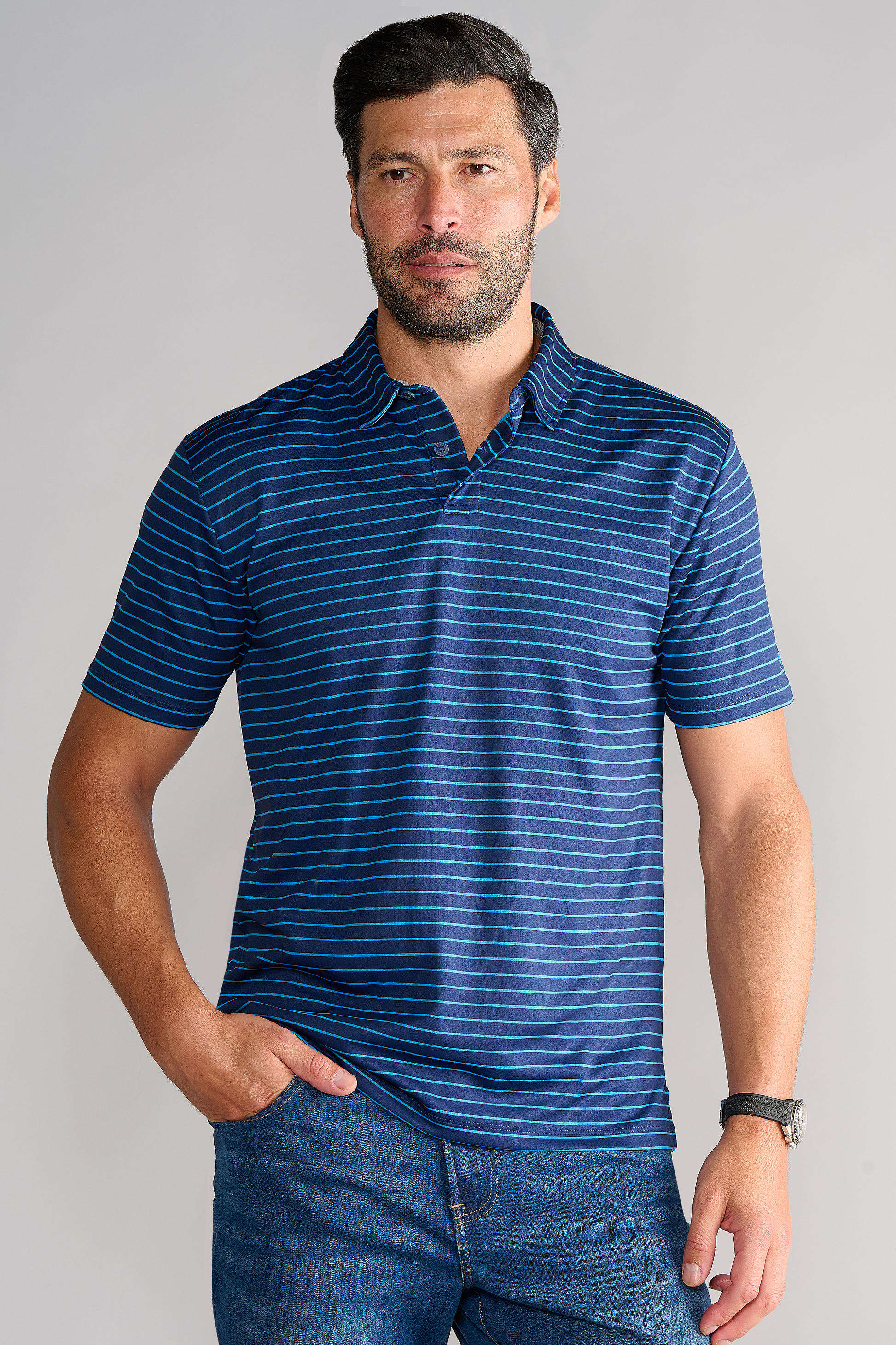 A man with short dark hair and a beard wears the Twillory Short Sleeve Polo Stripe in Navy Blue and blue jeans, standing against a light gray background with one hand in his pocket and a dark watch on his left wrist.