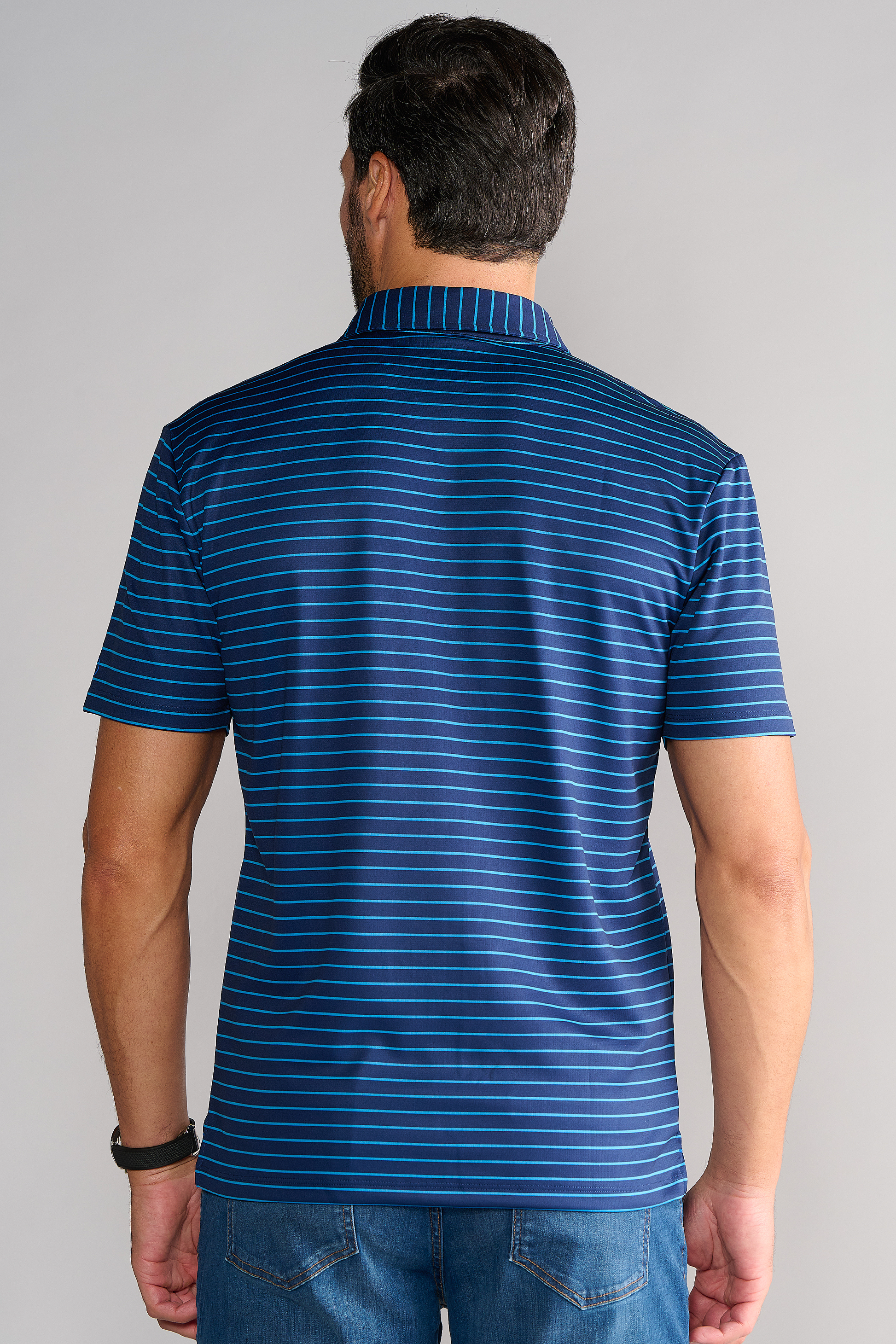 A man is seen from behind wearing the Twillory Short Sleeve Polo Stripe // Navy Blue, featuring thin light blue stripes and stretch fabric. He pairs it with jeans and a black watch, set against a plain light gray background.