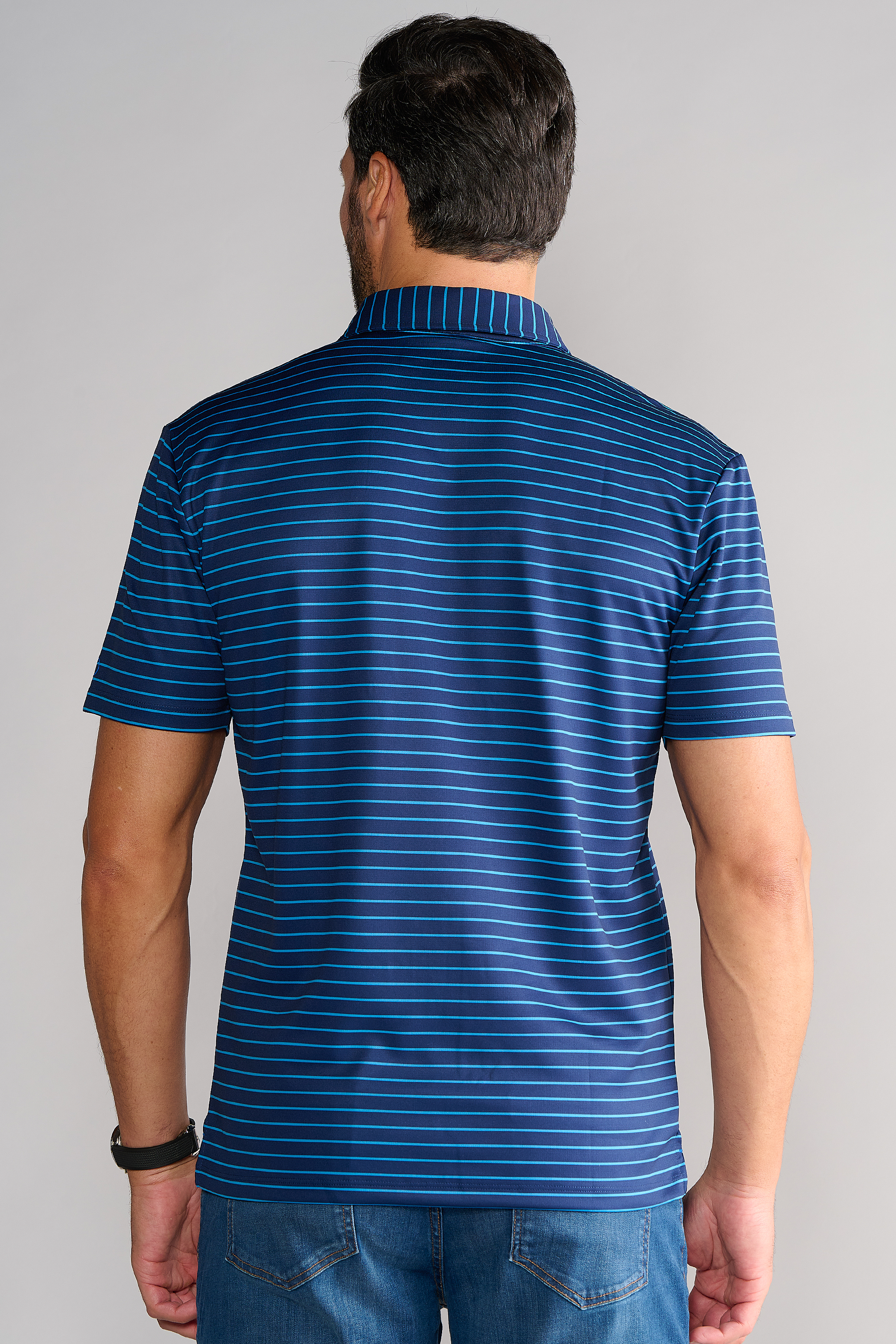 A man is seen from behind wearing the Twillory Short Sleeve Polo Stripe // Navy Blue, featuring thin light blue stripes and stretch fabric. He pairs it with jeans and a black watch, set against a plain light gray background.