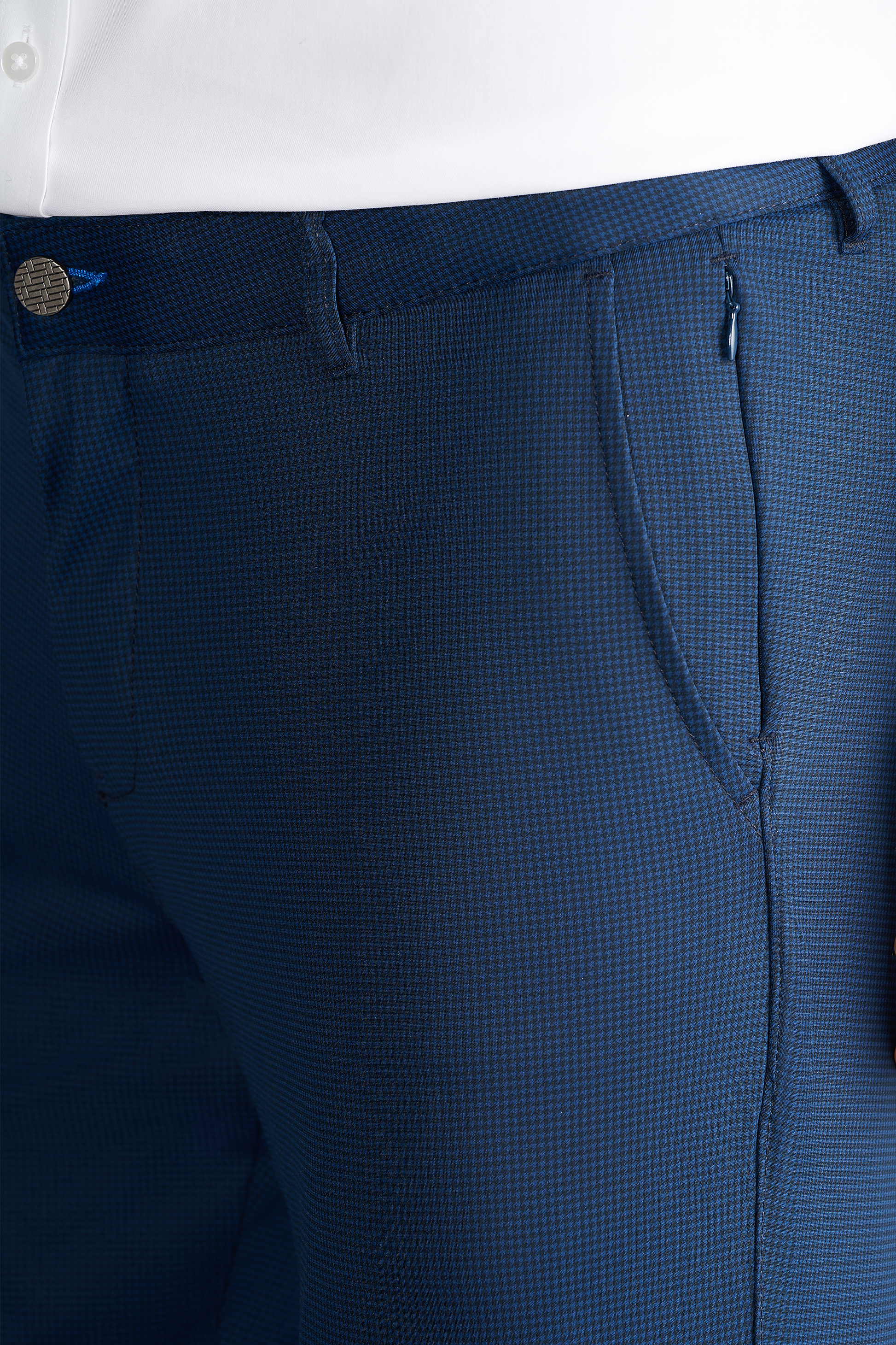 Close-up of a person wearing Twillory Performance Pants // Navy Houndstooth with front pockets, blue button, zipper, and visible stitching. Made from textured, non-iron performance fabric and paired with a white tucked-in shirt.