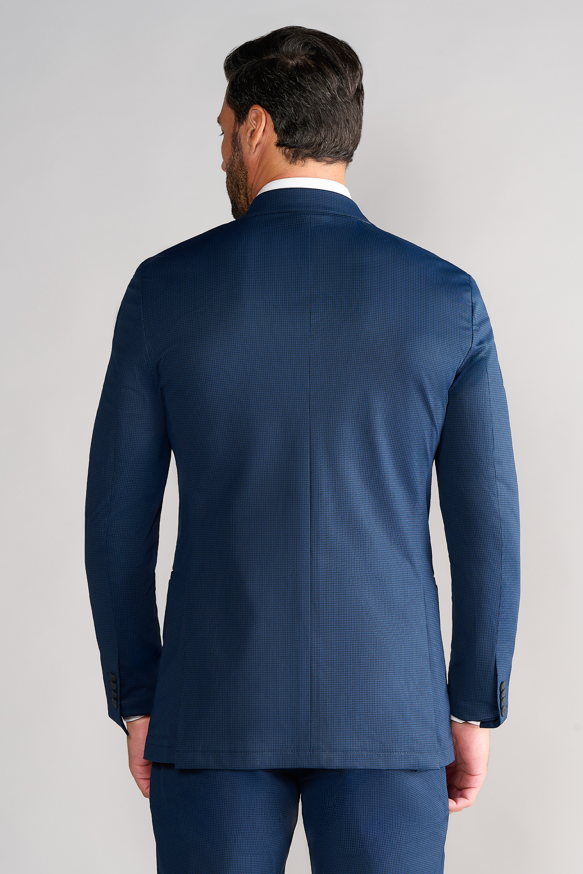 A man with dark hair is seen from behind, wearing Twillory's Performance Pants in Navy Houndstooth, featuring moisture-wicking, non-iron fabric. The plain light gray background highlights the pants' fit and textured design.