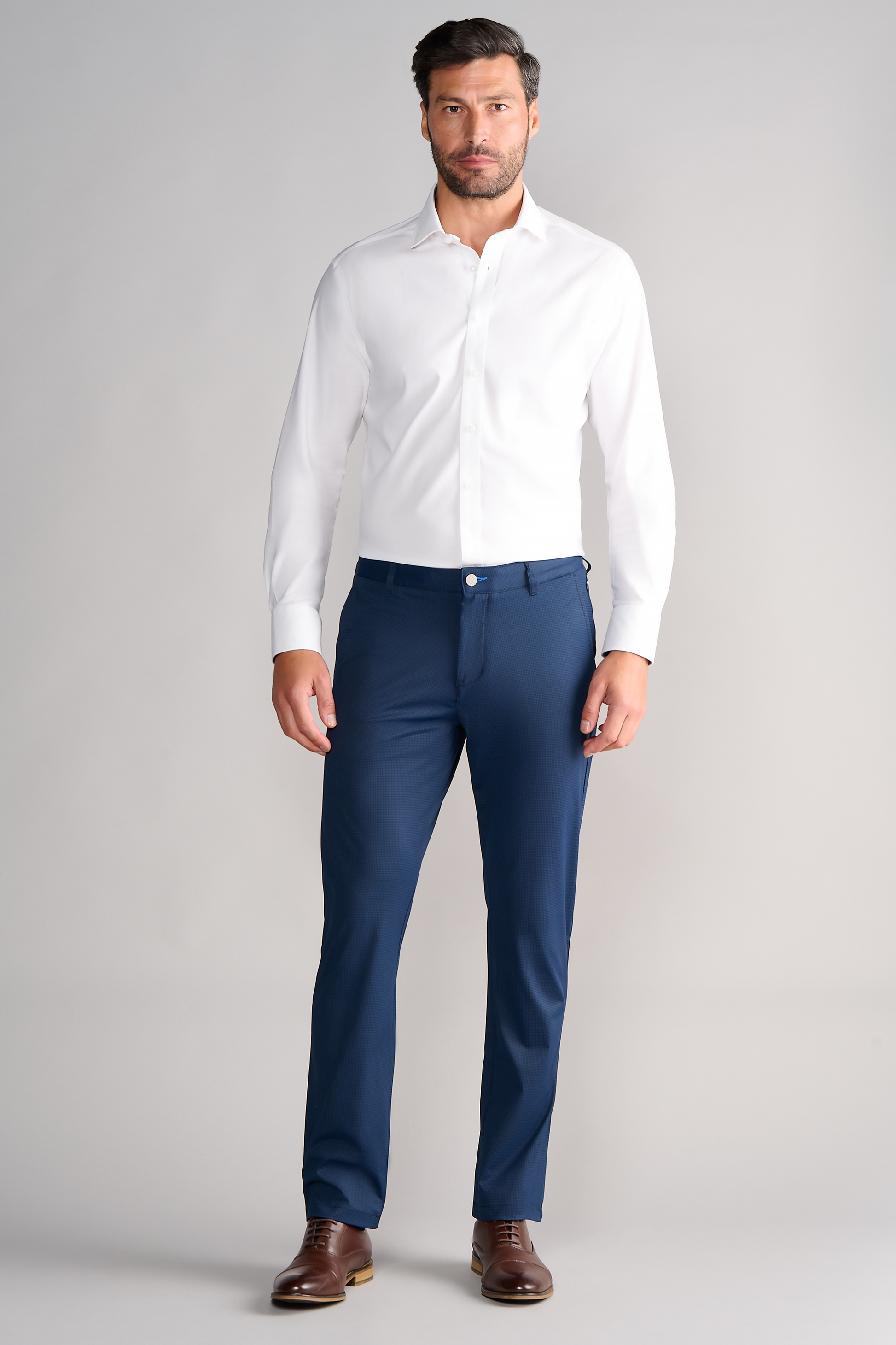 A man with short dark hair and a trimmed beard stands against a light gray background, wearing a fitted white dress shirt tucked into Twillory Performance Pants in Navy Houndstooth, paired with brown dress shoes. He looks forward neutrally, arms relaxed.