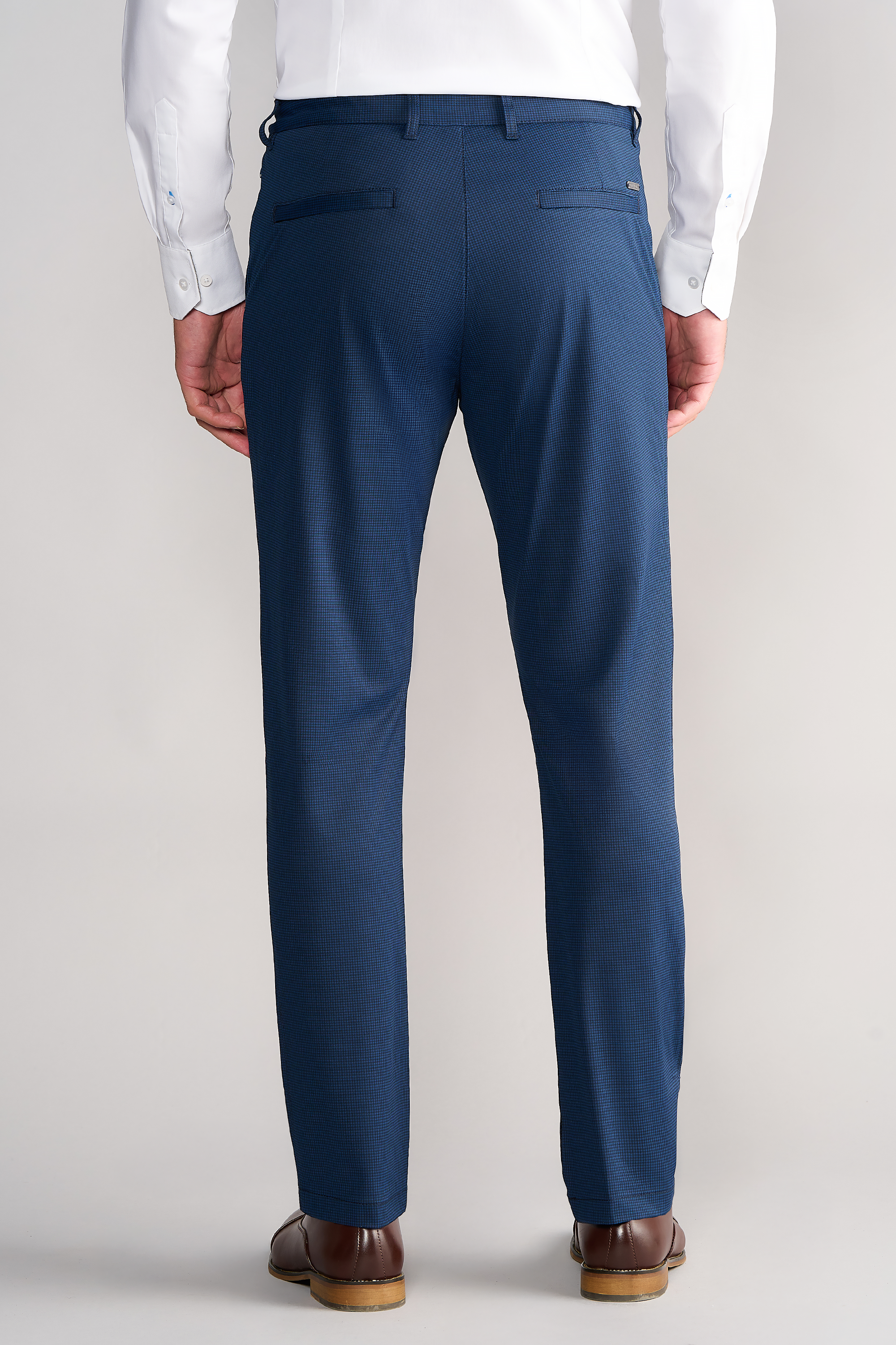 A man faces away, wearing a white dress shirt tucked into Twillory Performance Pants // Navy Houndstooth featuring 4-way stretch, non-iron fabric, and buttoned back pockets. He completes the look with brown dress shoes against a plain light background.