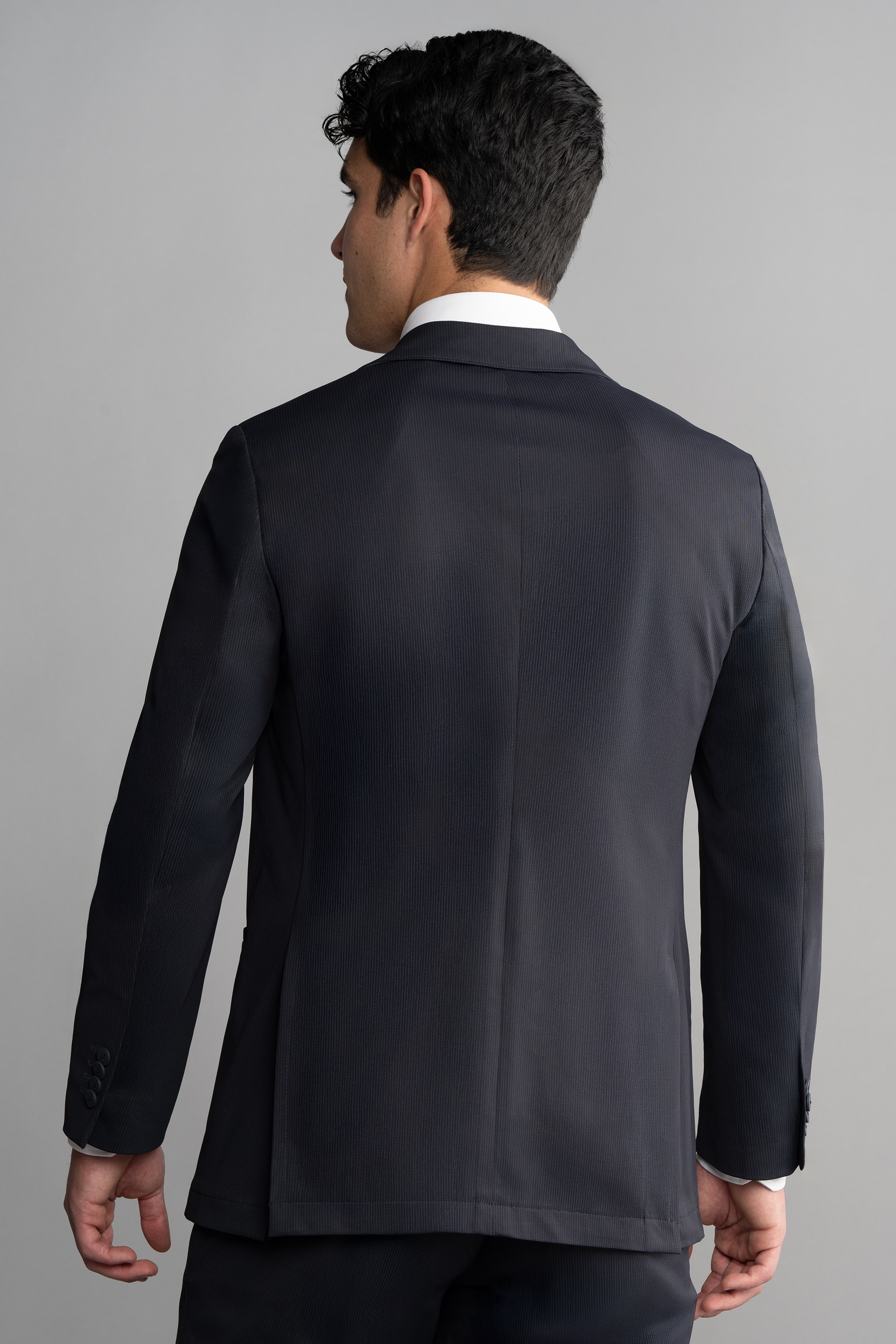 A man with short, dark, curly hair is seen from behind wearing Twillory’s Performance Blazer // Navy Pin Stripe and matching pants with a white dress shirt against a plain light gray background.