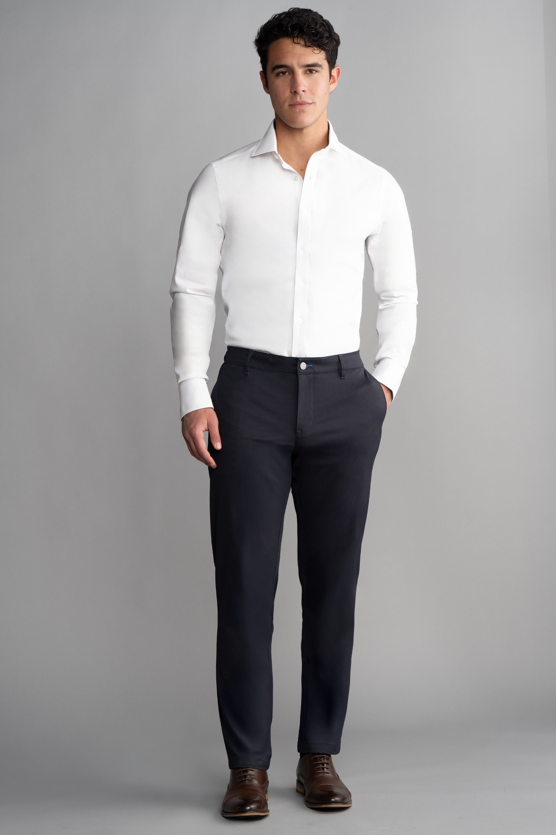 A man stands against a plain gray background, wearing a fitted white dress shirt and Twillory Performance Pants in Navy Pin Stripe, with brown leather shoes. His hands are in his pockets as he looks straight ahead with a neutral expression.