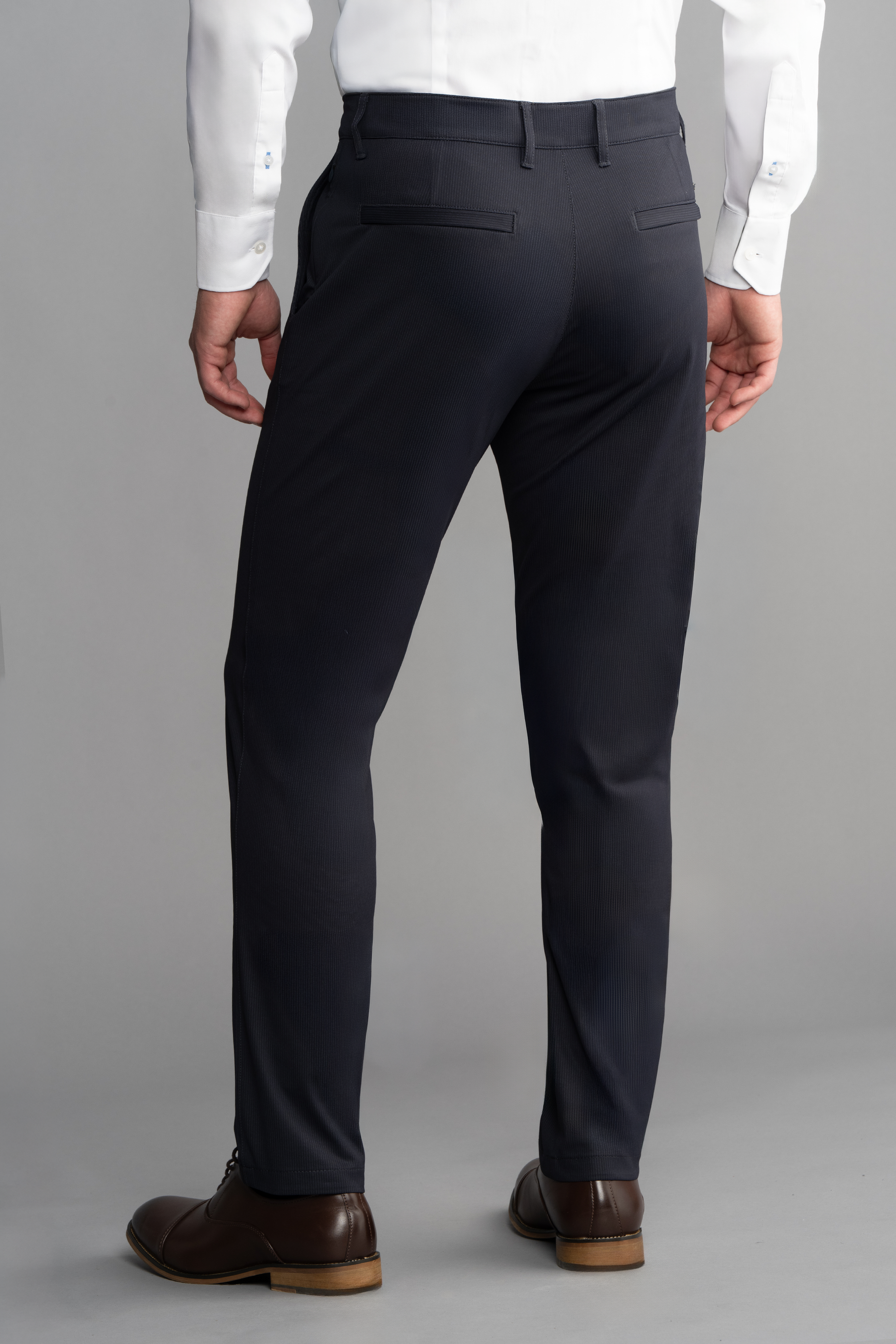 A man wearing Twillory Performance Pants in Navy Pin Stripe, a white dress shirt, and brown dress shoes stands facing away from the camera. The non-iron pants have two back pockets with button details. Background is plain light gray.