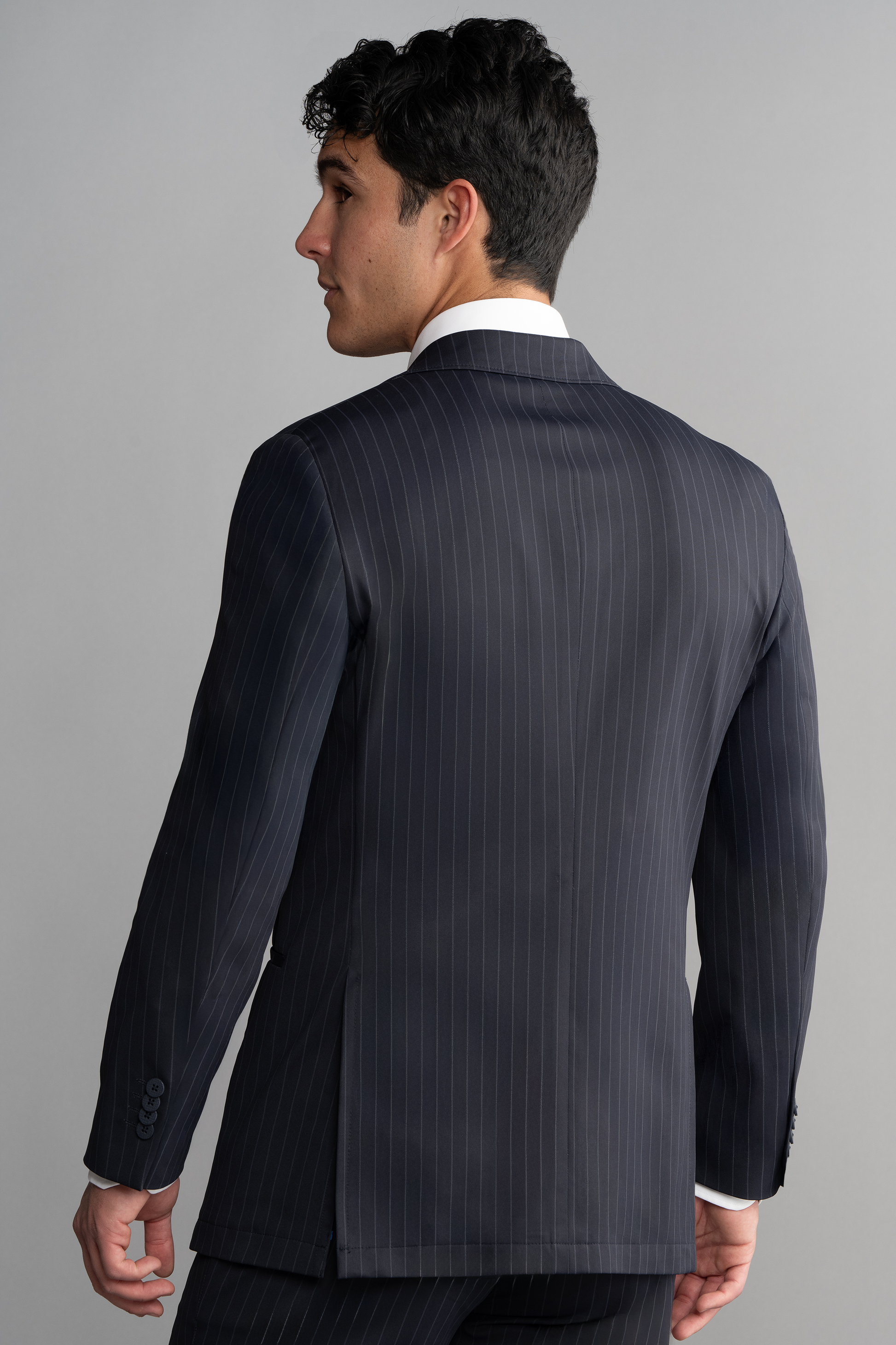 A man with short dark hair is seen from the back in a white shirt and Twillory Performance Blazer // Navy Stripe, featuring faint vertical stripes and a notch lapel. The plain light gray background shows his head turned slightly left.