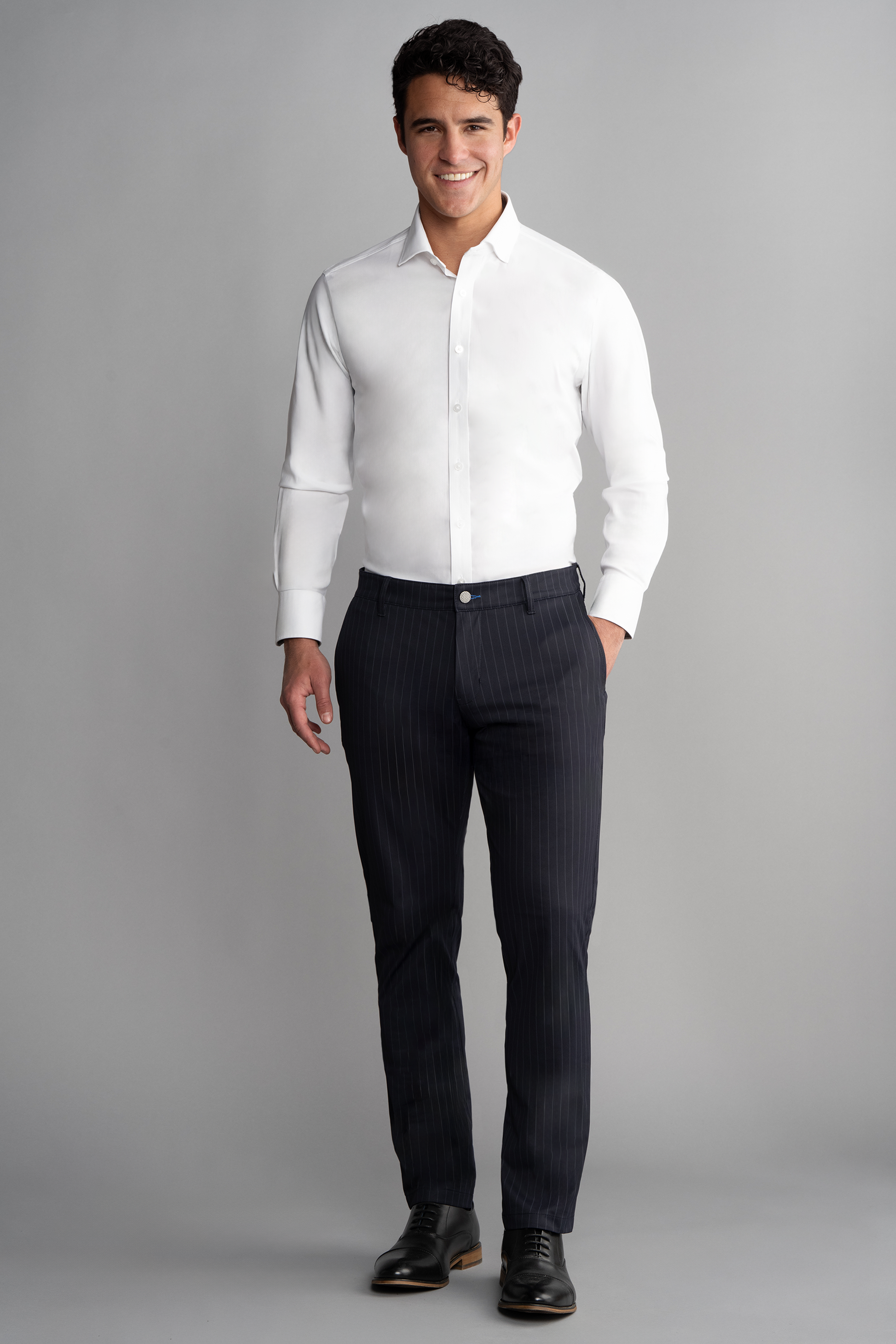 A man smiles against a gray backdrop, wearing a fitted white dress shirt, Twillory Performance Pants in Navy Stripe, and black dress shoes. His hands at his sides and short curly hair complete his professional, polished look.