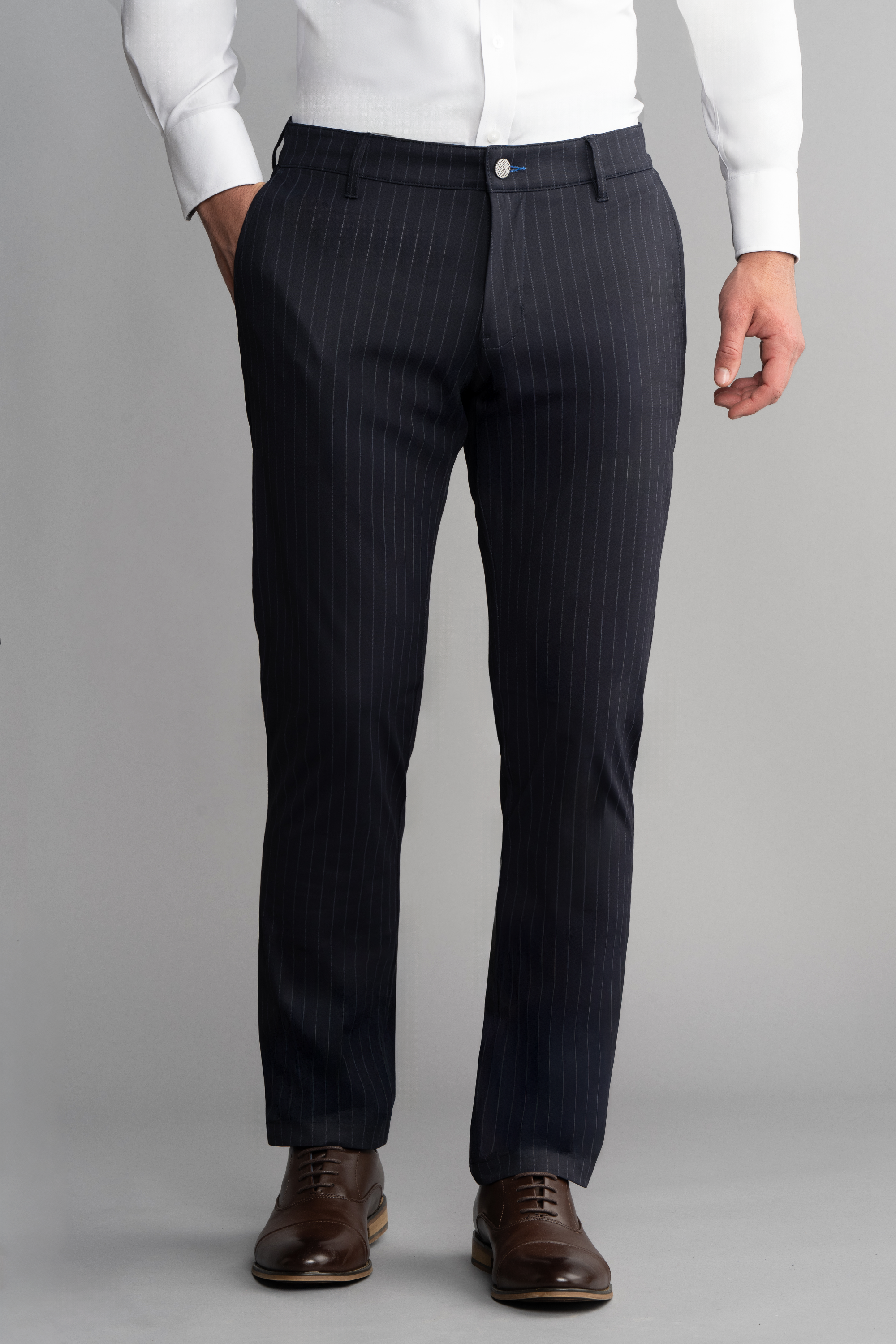 A man stands against a gray background, wearing a white dress shirt and Twillory Performance Pants // Navy Stripe—ideal for travel—with brown leather dress shoes. His left hand is in his pocket; only his body from shoulders down is visible.