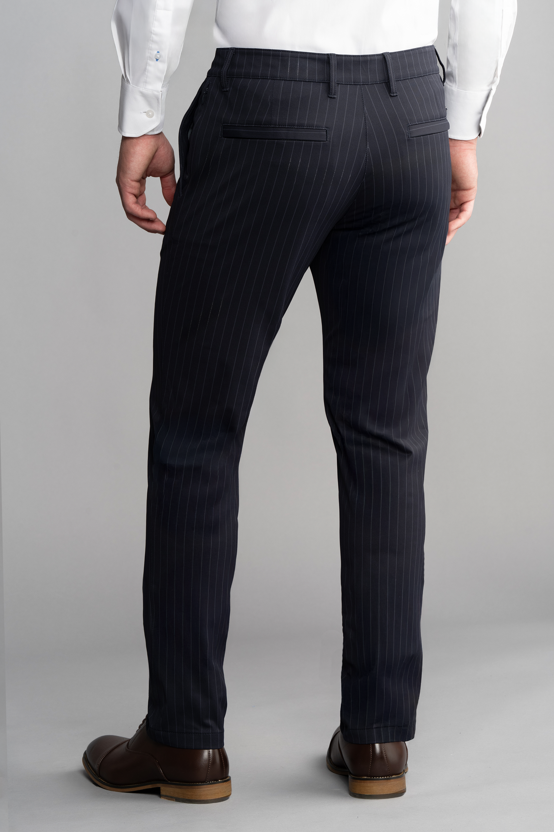 A man stands facing away, showcasing the back view of Twillory Performance Pants // Navy Stripe paired with a white shirt and brown dress shoes against a plain light gray background. The image highlights the lower half of his body.