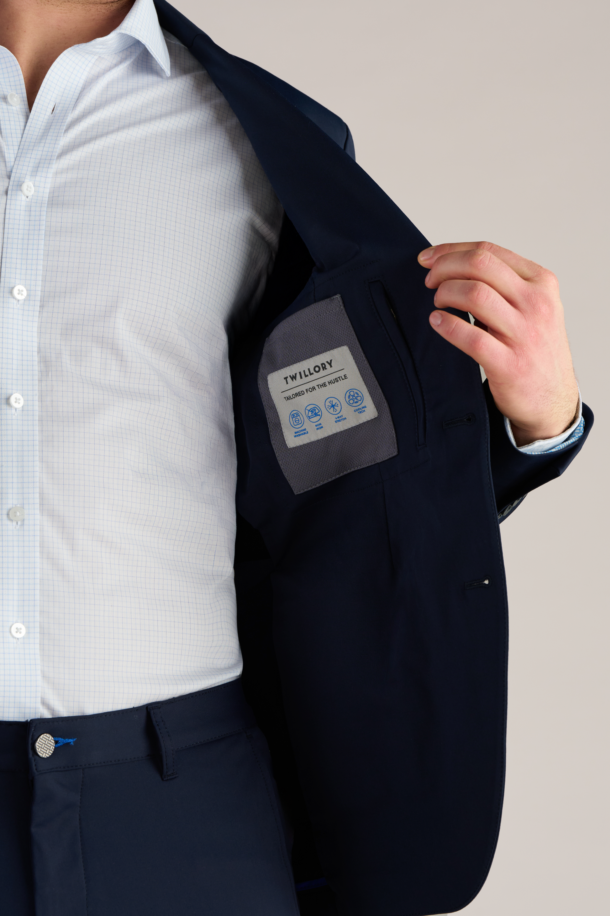 A man in a light blue checkered shirt and the Performance Blazer // Navy Blue by Twillory opens his jacket to show the inner pocket and Twillory label. Only his lower face is visible against a plain, neutral background.
