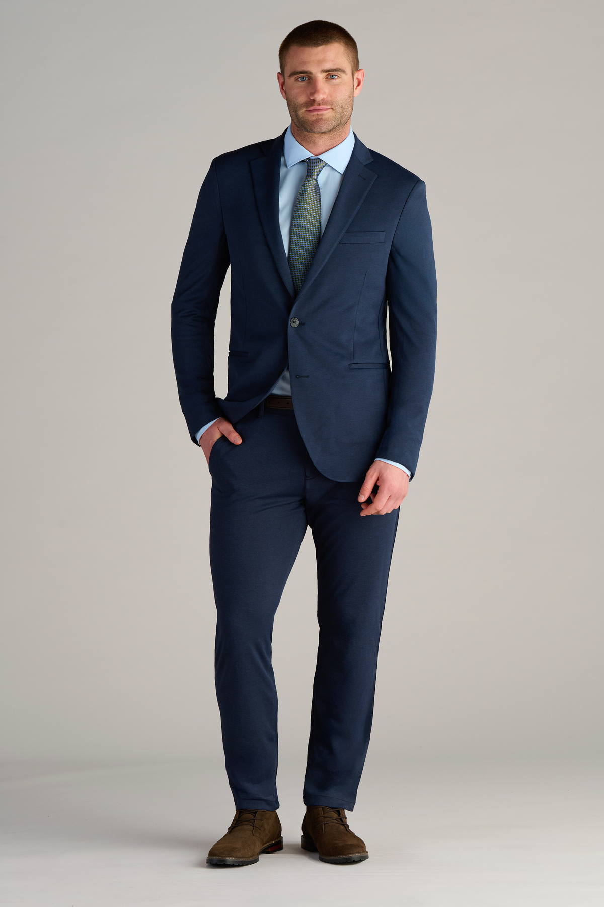 A man faces forward with one hand in his pocket, wearing a Twillory Circular Knit Blazer in Heather Navy over a light blue dress shirt, green patterned tie, brown belt, and brown suede shoes against a plain light-gray background.