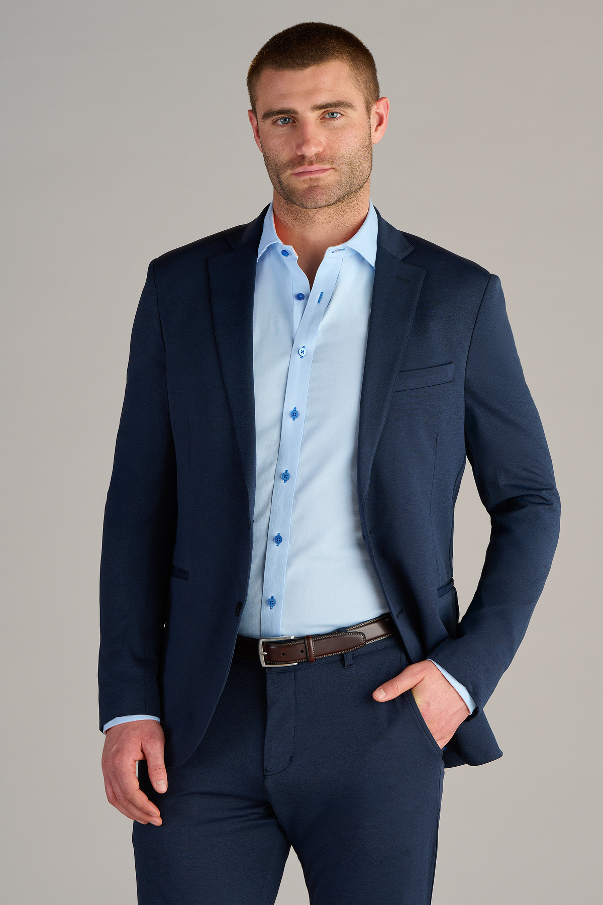 A man wears a Twillory Circular Knit Blazer // Heather Navy over a light blue dress shirt, buttoned up with no tie. He stands confidently with one hand in his pocket against a plain light gray background.