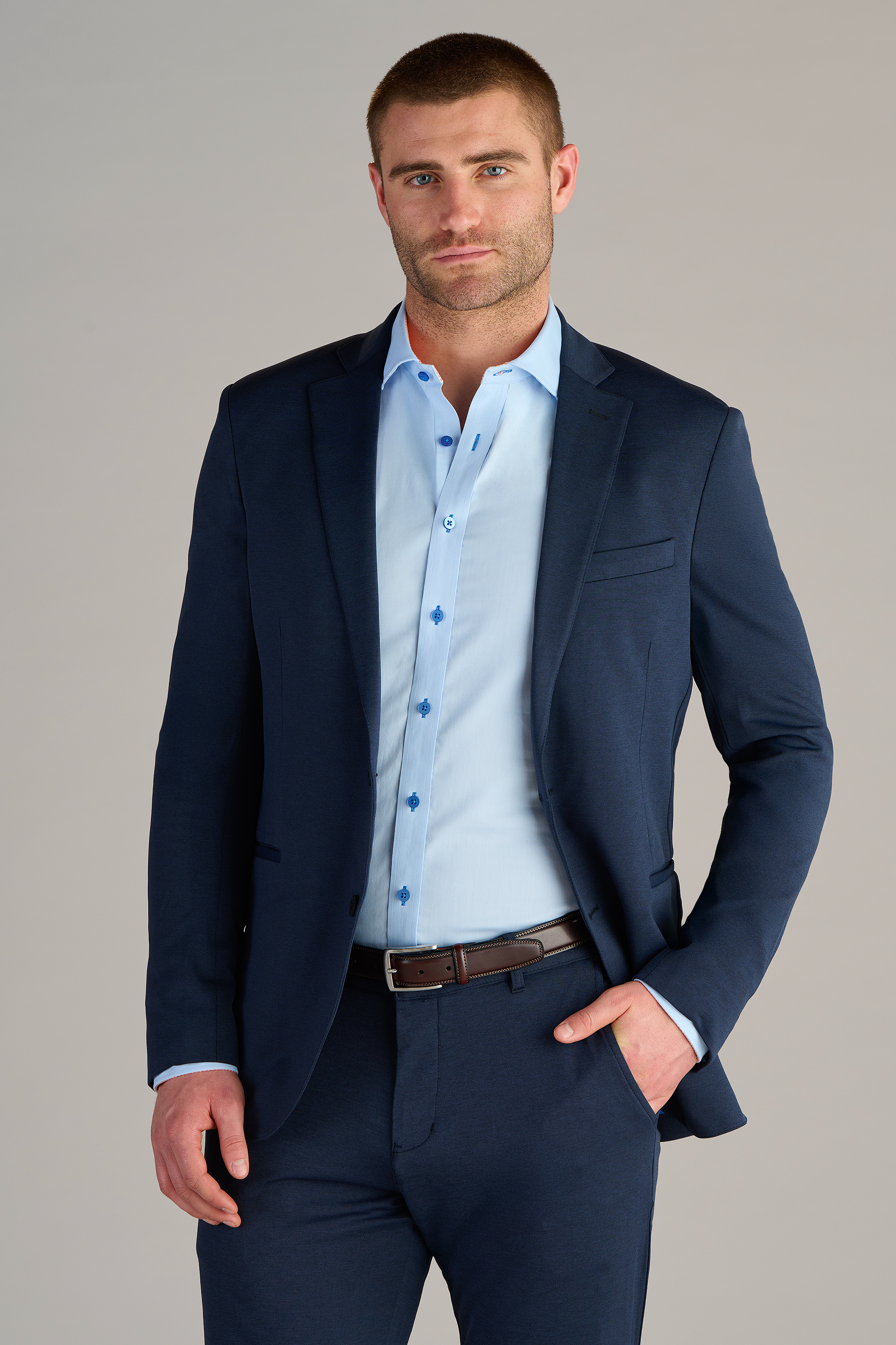A man wears a Twillory Circular Knit Blazer // Heather Navy over a light blue dress shirt, buttoned up with no tie. He stands confidently with one hand in his pocket against a plain light gray background.