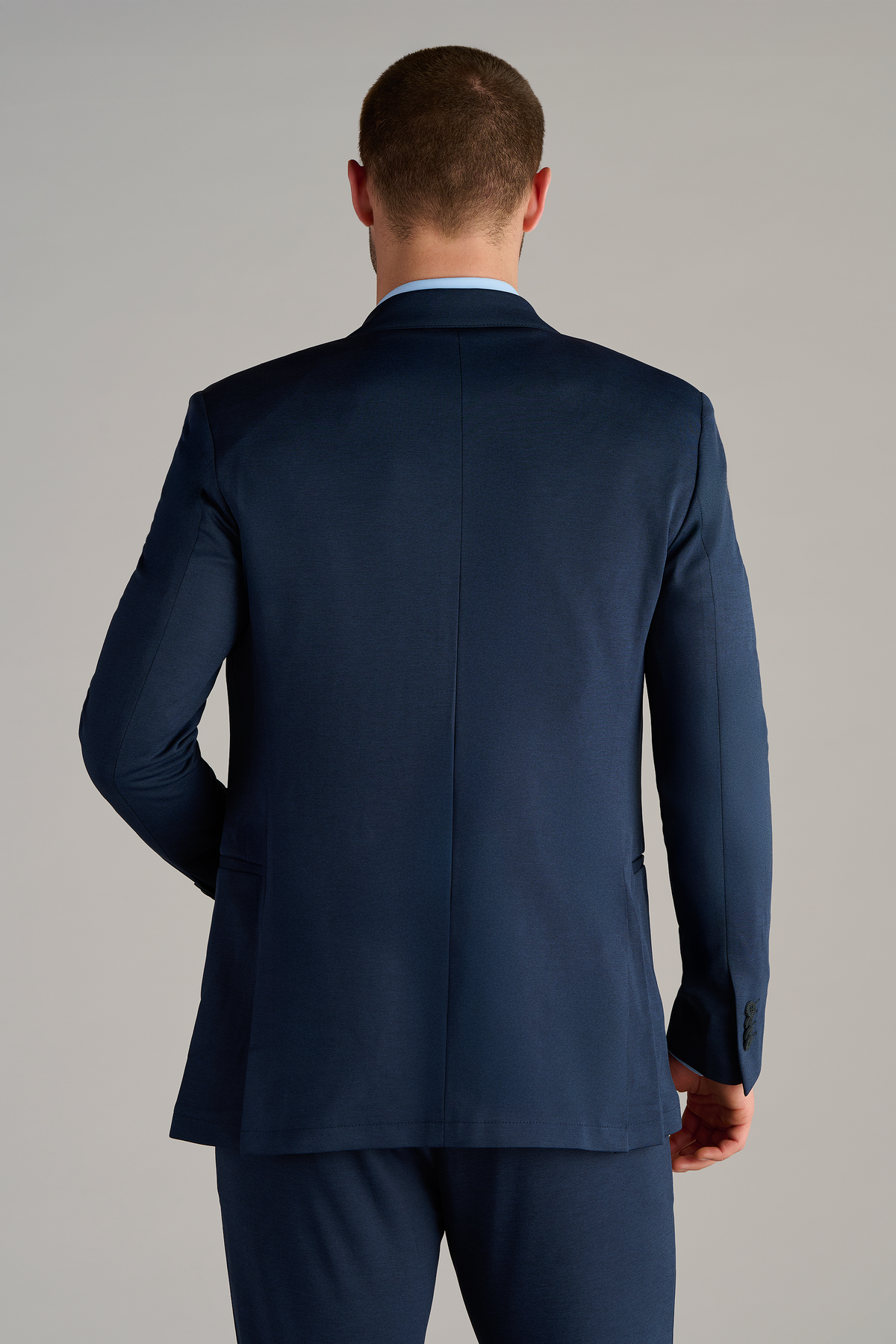 A man with short brown hair is seen from behind, wearing a Twillory Circular Knit Blazer in Heather Navy with matching trousers. His right arm is slightly bent, and the smooth-fitting jacket features a visible center seam against a light gray background.