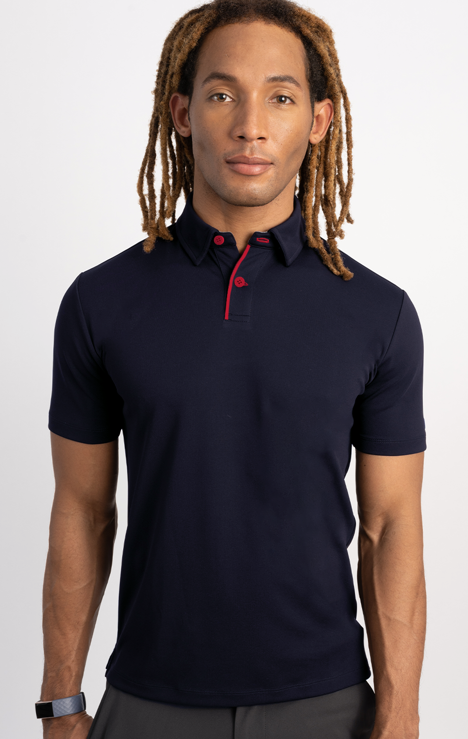 A person with long, light brown dreadlocks faces forward, wearing Twillory's Short Sleeve Polo Contrast // Navy Red with a red-lined placket and matching red buttons. The background is plain white; they also have a black wristband.