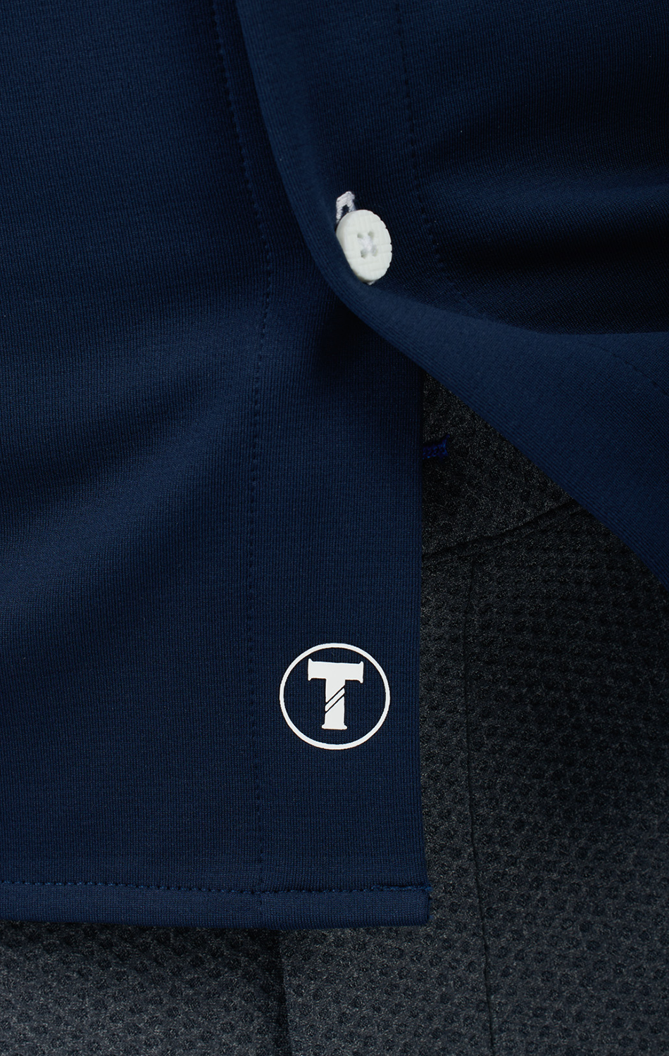 A close-up of Twillory's Long Sleeve Button Down Polo Contrast in Navy White, featuring a white button and stitched logo near the hem. The bold T-in-circle stands out against the textured gray background for striking contrast.