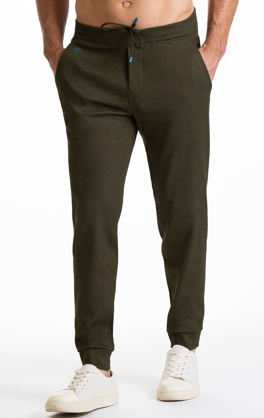 A person wears Twillory's Athlounger Jogger in olive green, featuring a tapered fit, drawstring waist, and side-zip pockets. Paired with white sneakers, only the lower torso and legs are visible against a white background.