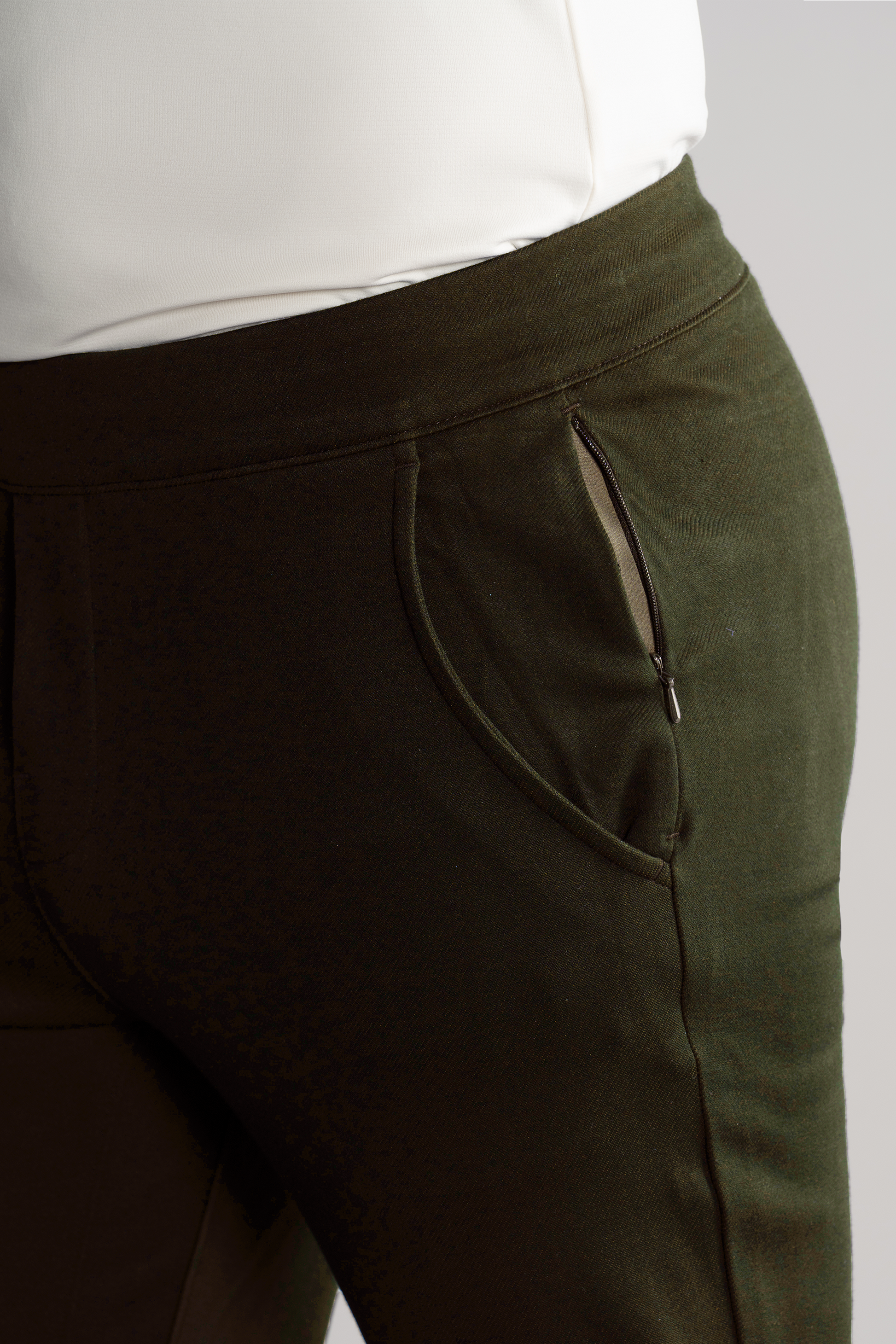 A close-up of a person wearing Twillory's Athlounger Jogger in dark green, featuring a silver side-zip pocket on the right. The tapered fit is styled with a tucked-in white top against a neutral gray background.