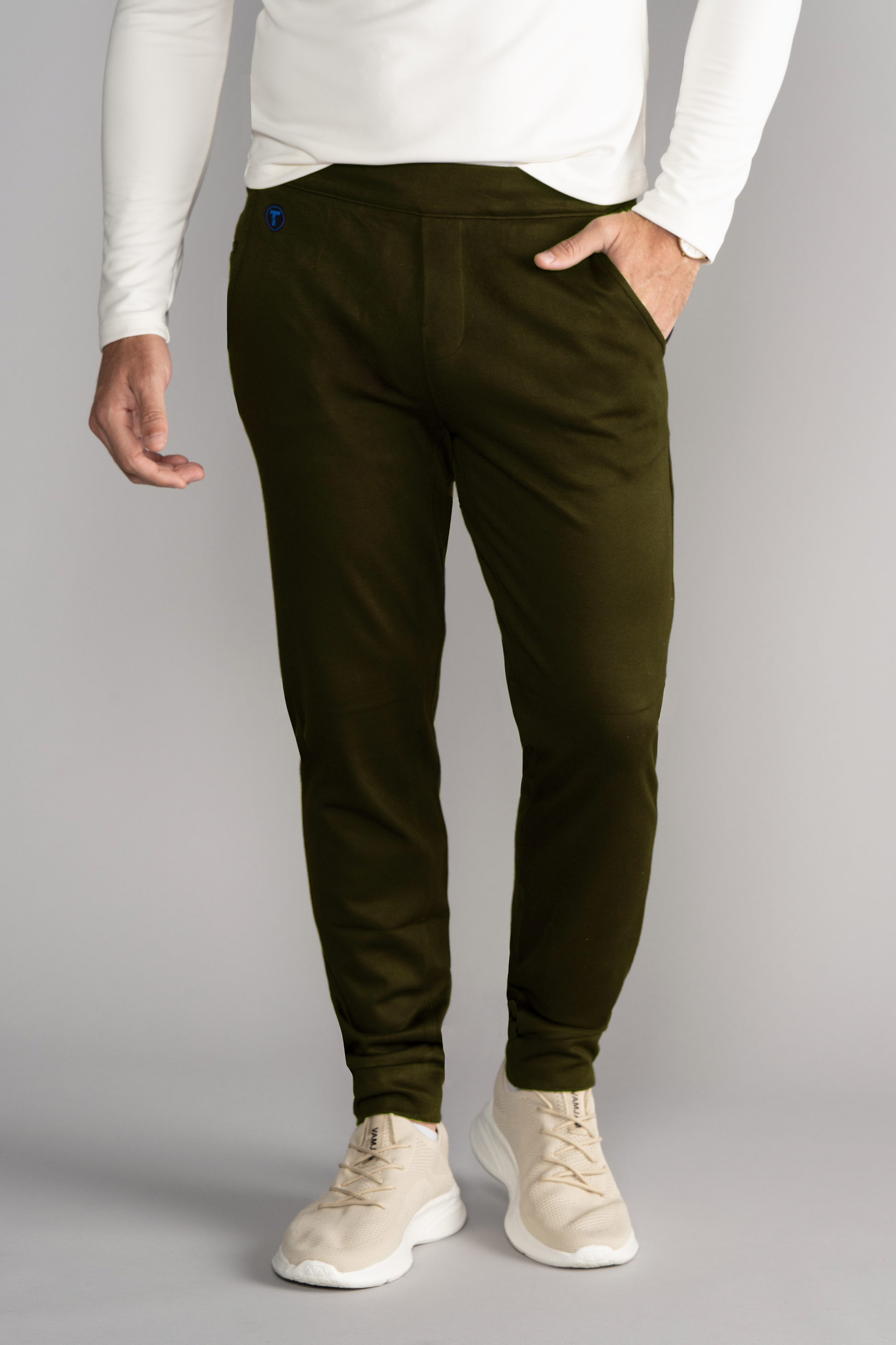 A person models Twillory's Athlounger Jogger in olive green, paired with a white long-sleeve shirt and light beige sneakers against a neutral backdrop. One hand is in their pocket; the other arm hangs relaxed, with their head out of frame.