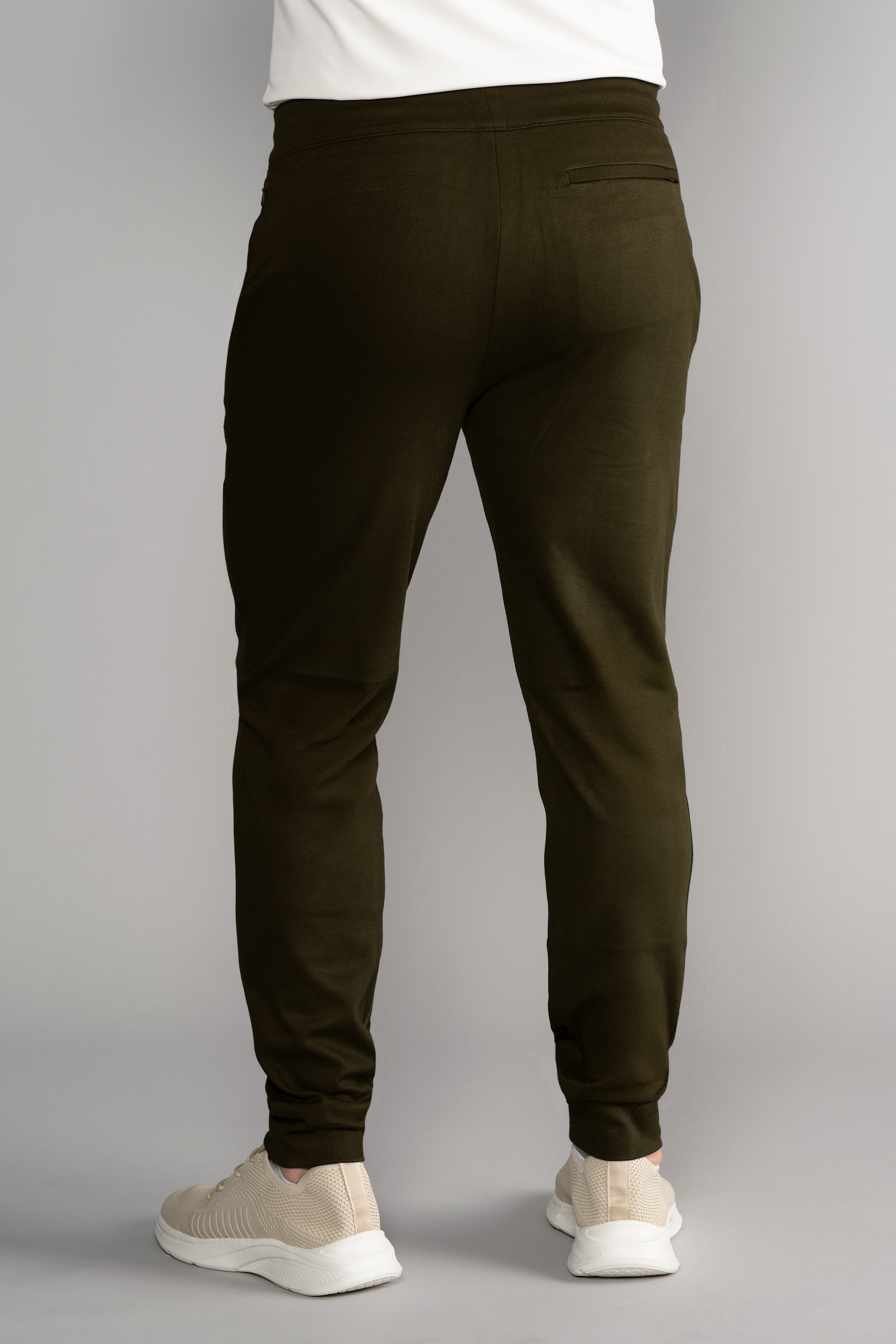A person wearing Twillory's Athlounger Jogger in olive green, featuring a tapered fit and a right back pocket, with white sneakers, stands facing away from the camera against a plain gray background. Only the lower back and legs are visible.