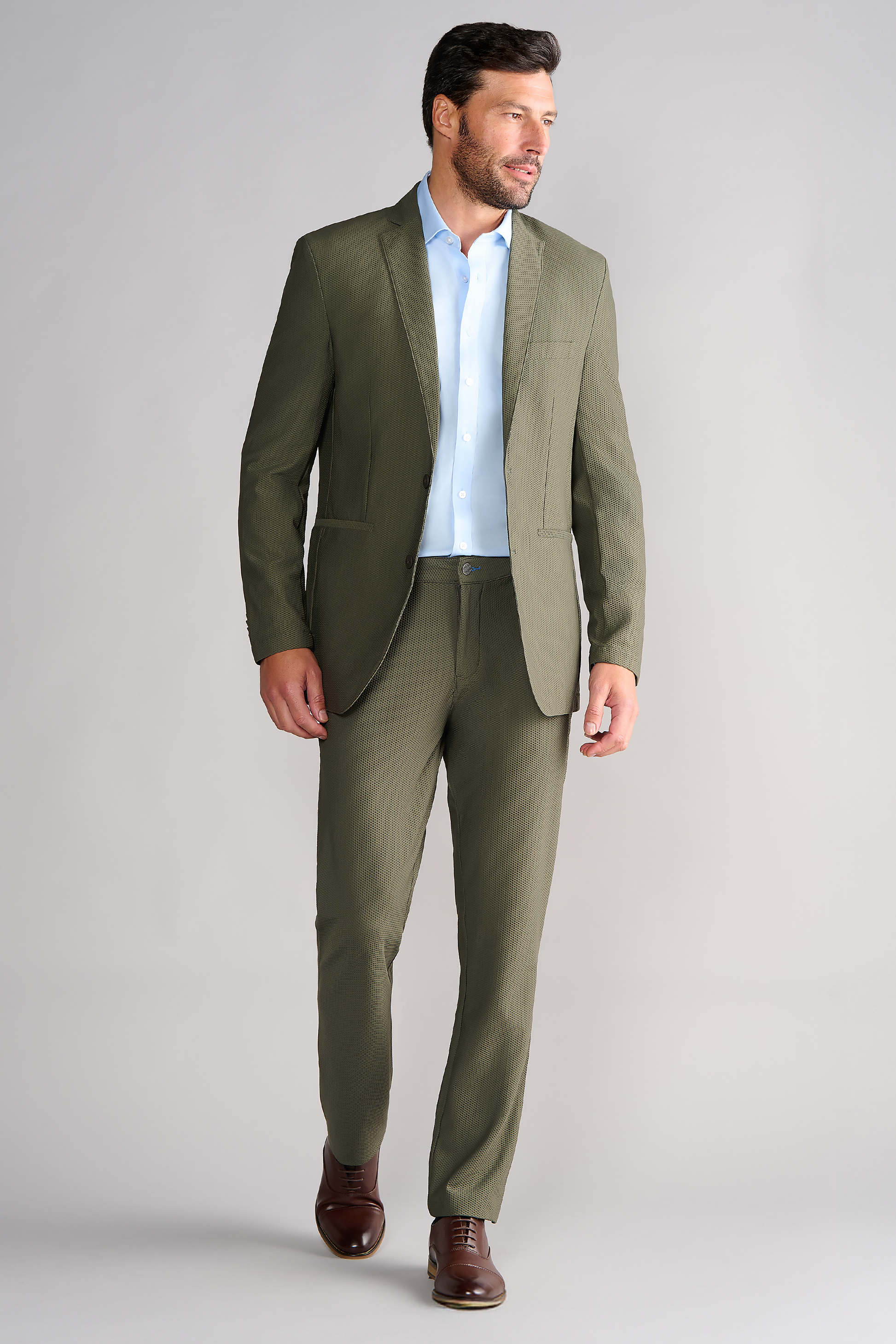 A man with short dark hair and a trimmed beard walks against a gray background, wearing the Twillory AIR Blazer in Olive—crafted from 360° Airflow Fabric—paired with a light blue shirt and brown dress shoes. He looks left with a neutral expression.