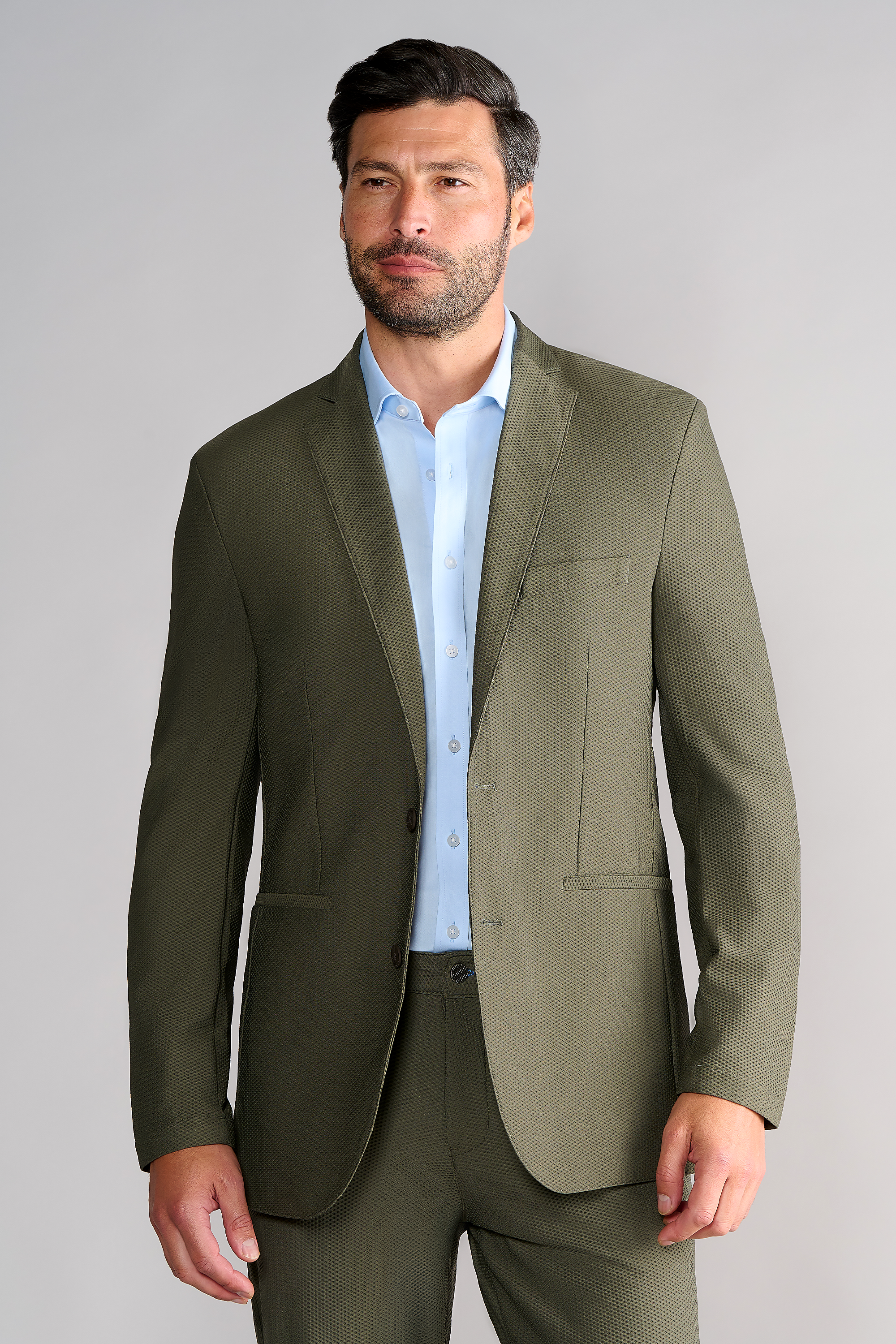 A man with short dark hair and a trimmed beard wears the Twillory AIR Blazer in Olive over a light blue dress shirt, standing against a plain light gray background. He looks slightly left with a neutral expression, his right hand relaxed by his side.