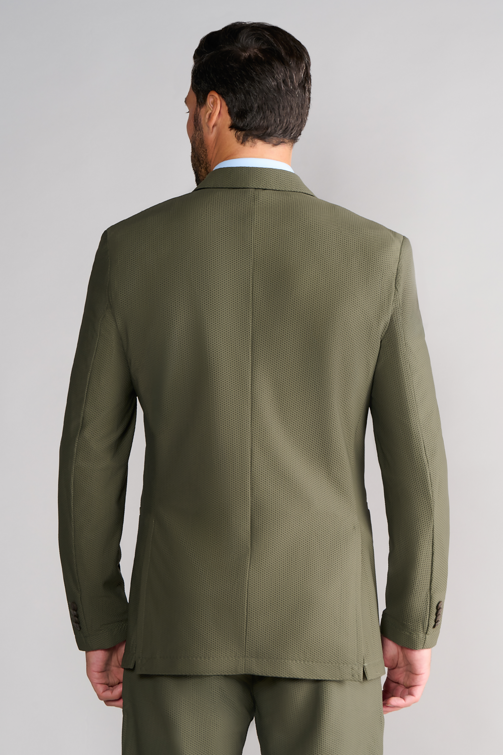 A man is seen from behind wearing Twillory’s AIR Blazer // Olive, tailored from 360° Airflow Fabric with a subtle texture and dual vent slits, paired with matching pants, standing against a light gray background.