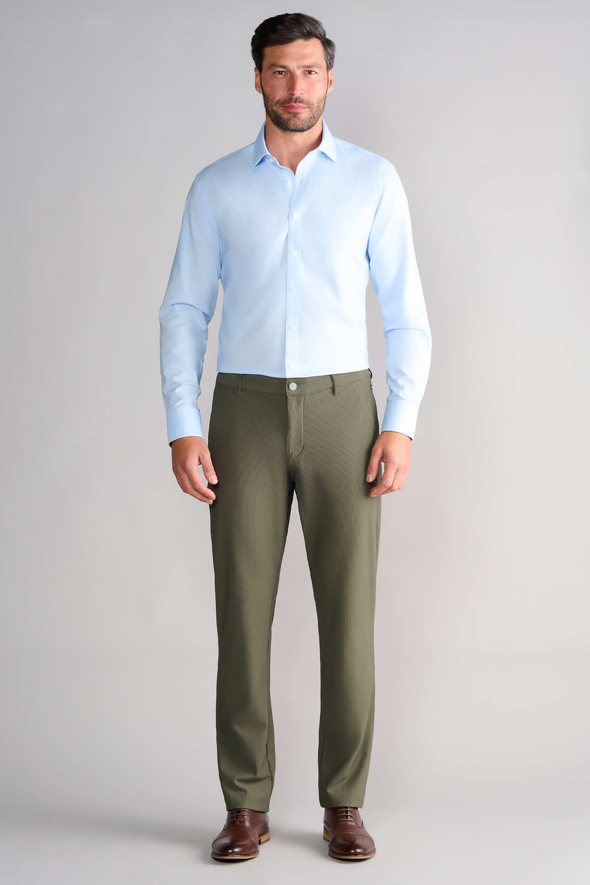 A man with short dark hair and a trimmed beard stands against a light gray background wearing Twillory AIR Pants in Olive, paired with a light blue dress shirt and brown shoes. The pants feature 360° Airflow Fabric for comfort.