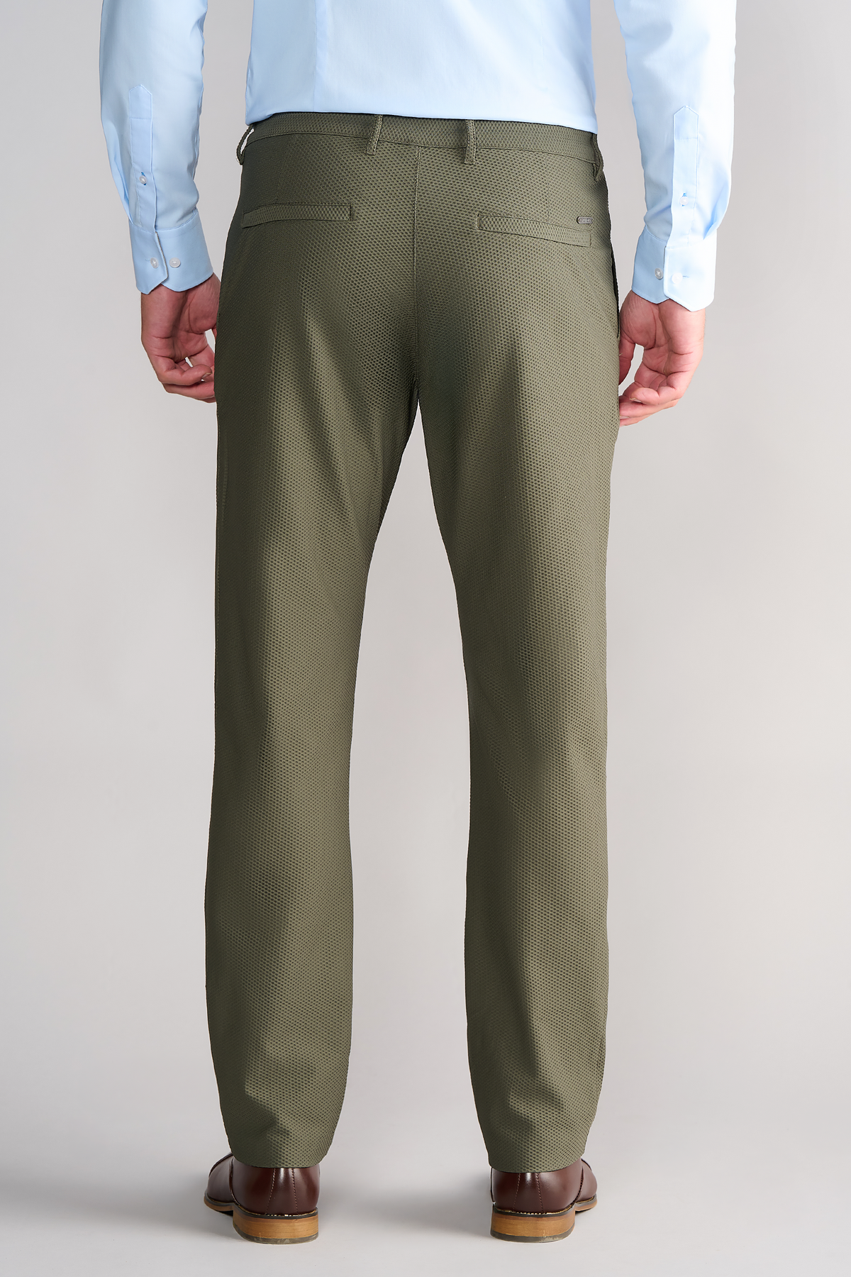 A man faces away in Twillory AIR Pants // Olive, featuring 360° Airflow Fabric, subtle vertical texture, and two buttoned rear pockets. He pairs them with a light blue shirt and brown dress shoes against a plain light grey background.
