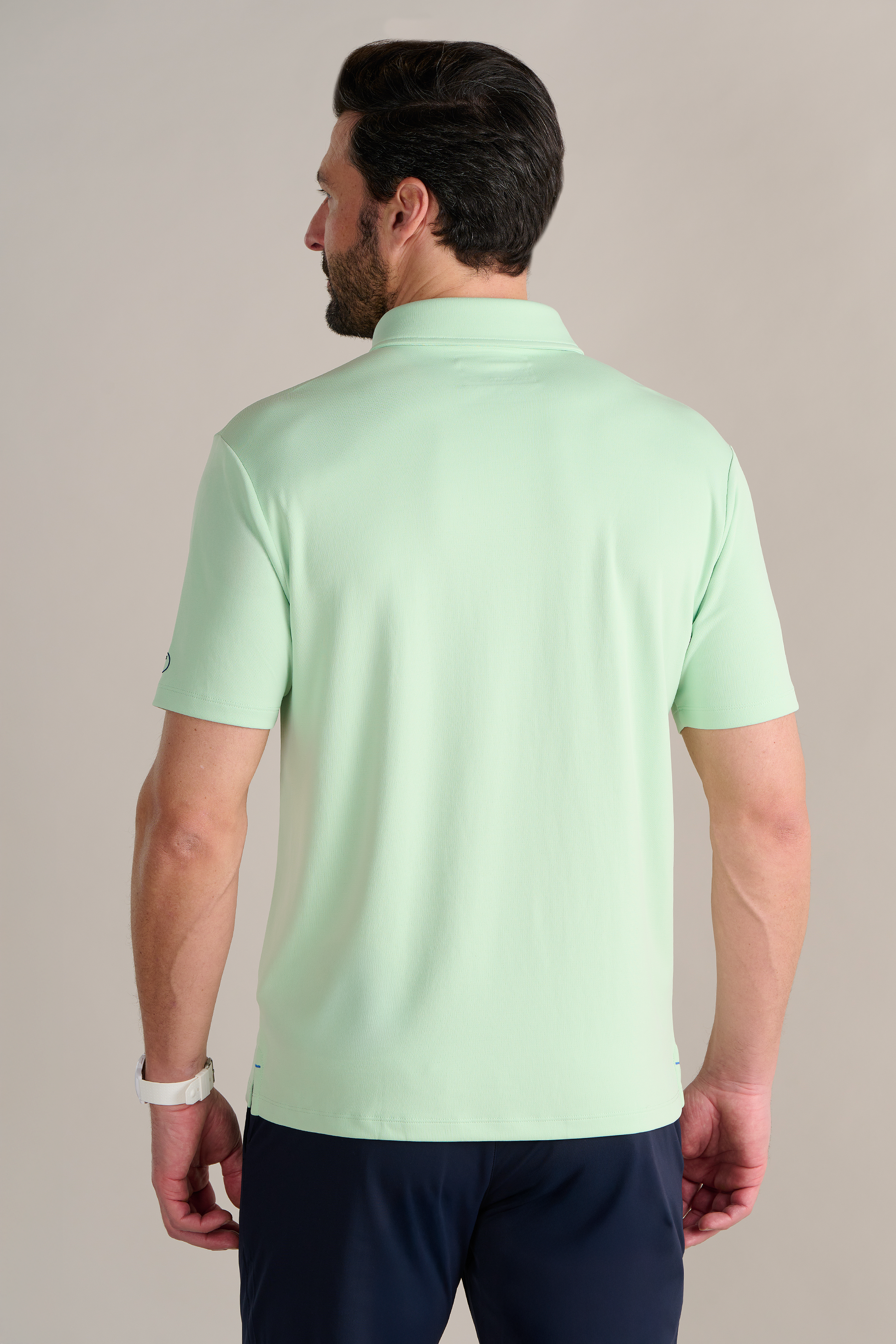 A man with short dark hair and a beard is seen from behind, wearing the Twillory Short Sleeve Polo Solid in Pastel Green and dark pants. He sports a white watch on his left wrist against a plain, light-colored background.
