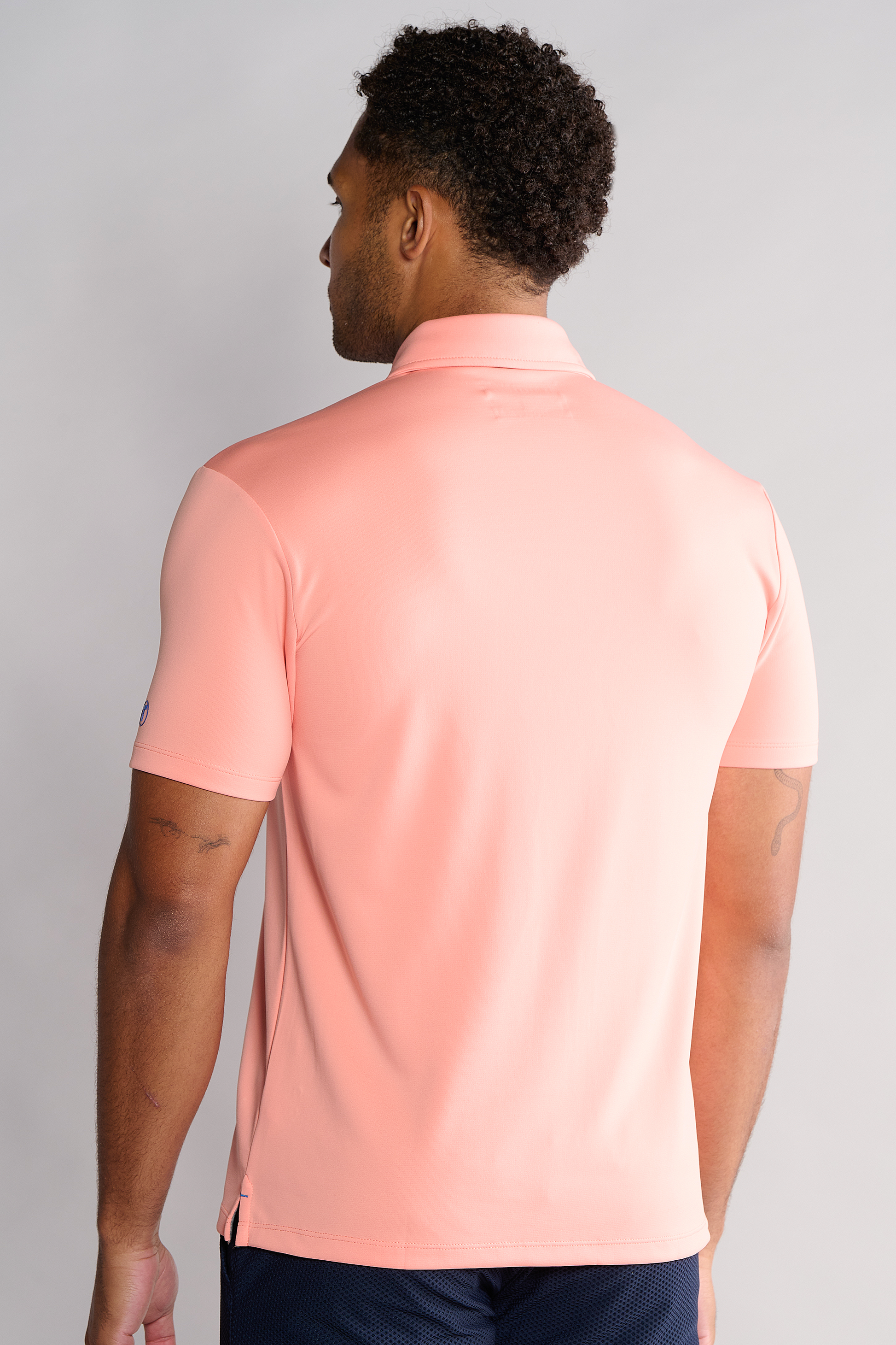 A man with short curly hair faces away, wearing the Twillory Short Sleeve Polo Solid in Peach and dark shorts. The plain, light background highlights the back view of this comfortable performance polo with a dress collar.