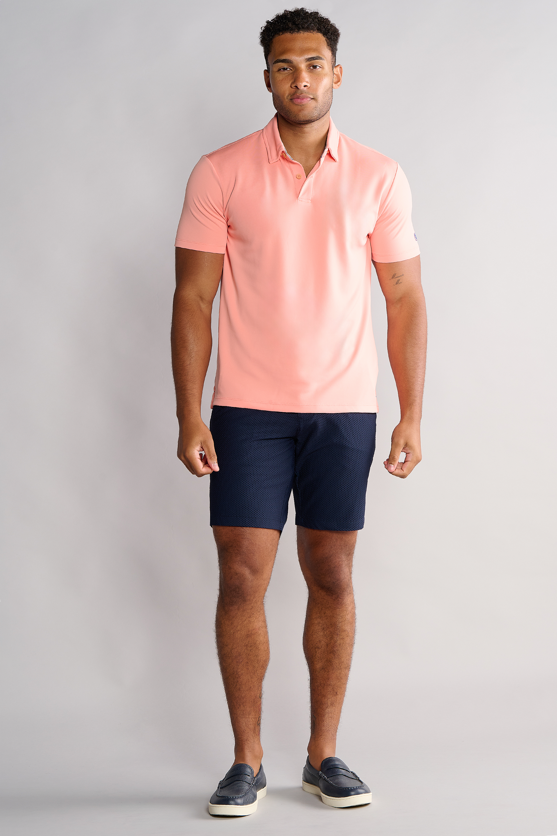 A man with short curly hair and a trimmed beard stands against a plain background, wearing the Twillory Short Sleeve Polo Solid in Peach, paired with navy shorts and dark slip-on shoes, arms relaxed by his sides.