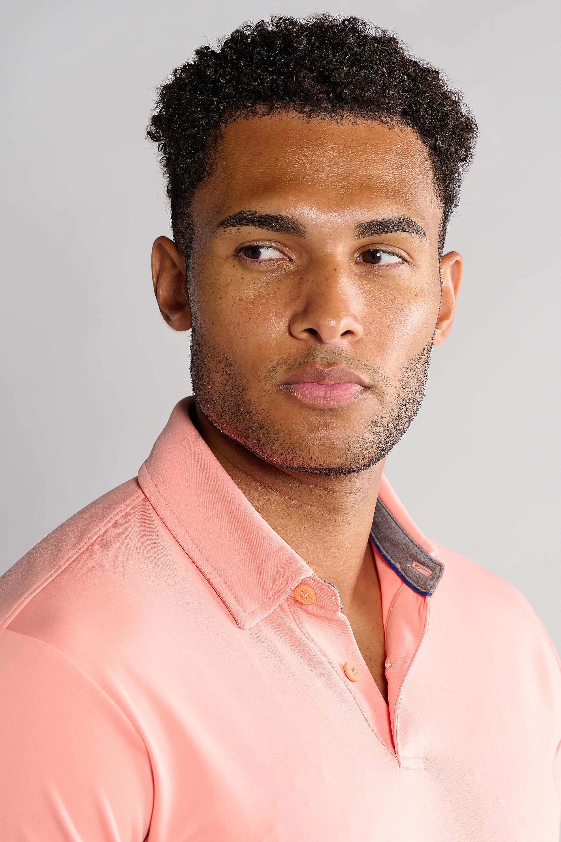 A man with short, curly hair and trimmed facial hair wears the Twillory Short Sleeve Polo Solid in Peach. He faces left with a neutral expression against a plain light gray background, showing the shirt’s dress collar and dark inner lining.