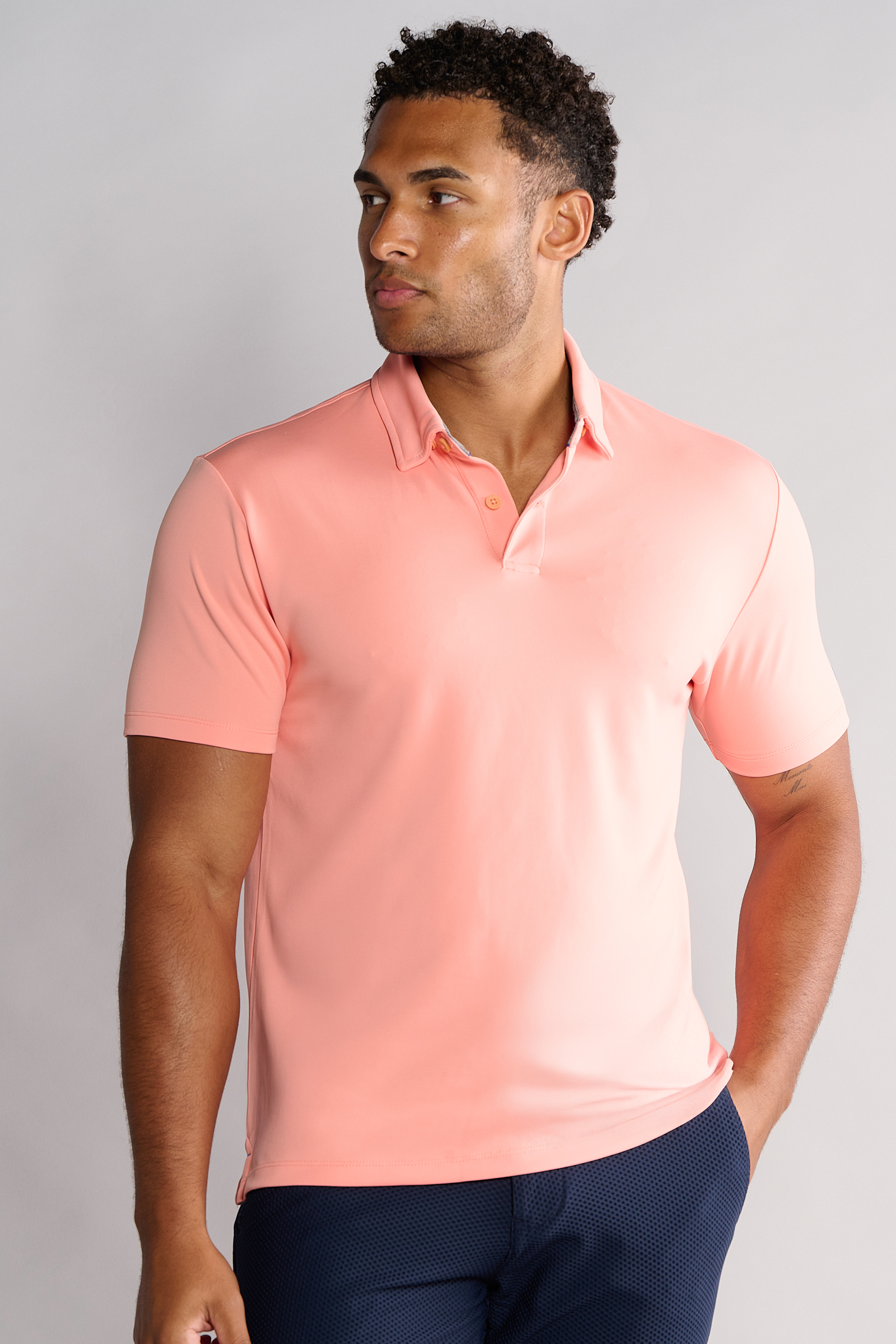 A man with short curly hair stands against a light gray background wearing the Twillory Short Sleeve Polo Solid in Peach and dark navy pants, looking to his left with a neutral expression and one hand in his pocket.