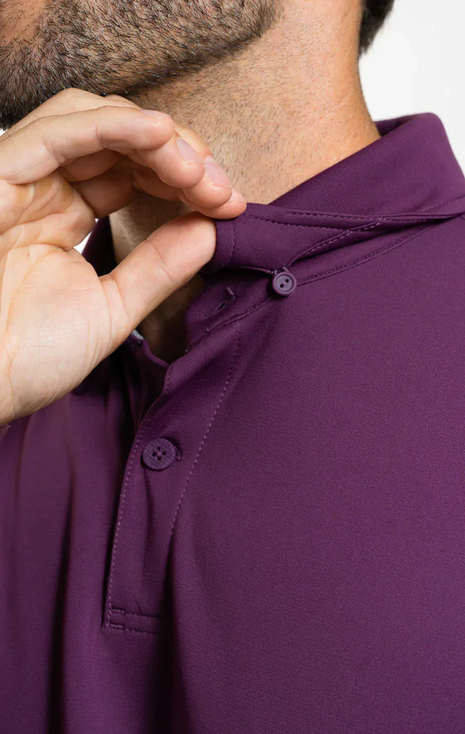 A close-up of a person wearing the Twillory Short Sleeve Polo Solid in Plum, adjusting the dress collar with their left hand near a small button. The lower part of their bearded face appears against a plain light background.